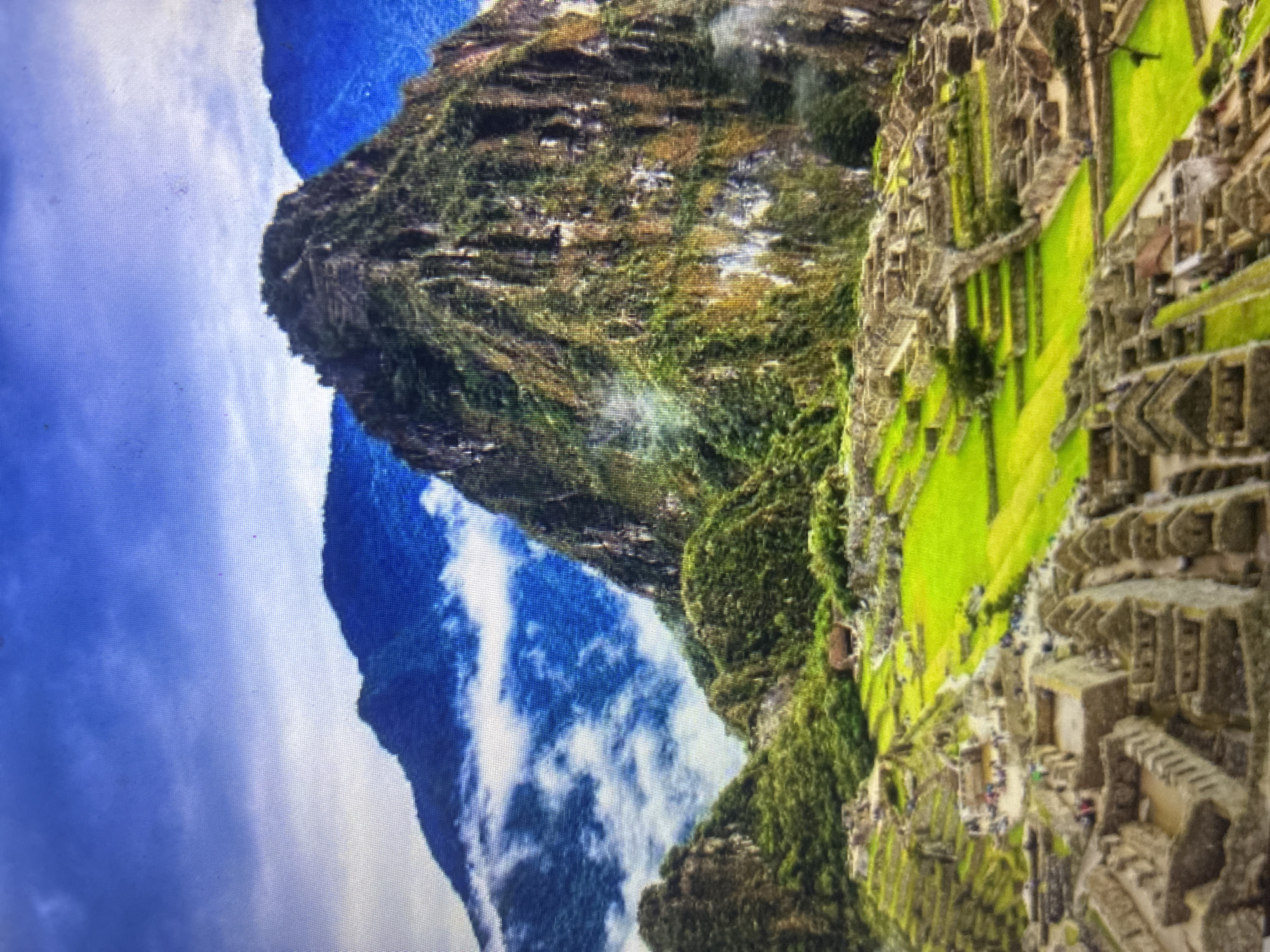 The image displays a panoramic view of an expansive ancient city, strongly resembling Machu Picchu, situated high in a mountainous, verdant landscape. The scene is dominated by intricate stone terracing and numerous well-preserved stone structures, characteristic of Inca architecture, built into the steep slopes of a mountain. Lush green vegetation covers the surrounding terrain, with towering peaks visible in the background, some partially enveloped by white clouds and mist. The sky above shows patches of blue interspersed with white, fluffy clouds, suggesting daytime conditions. Small, indistinct figures, likely visitors or tourists, can be observed moving along the pathways and exploring the ruins. The photograph itself appears to be an image captured from a digital screen, displaying a noticeable grid-like pixel pattern, along with some slight blurring and potential dust artifacts on the screen surface. There is no visible text in the image. Although the provided context states Asenovgrad, Bulgaria, the visual evidence overwhelmingly identifies the depicted location as the historic sanctuary of Machu Picchu in Peru.