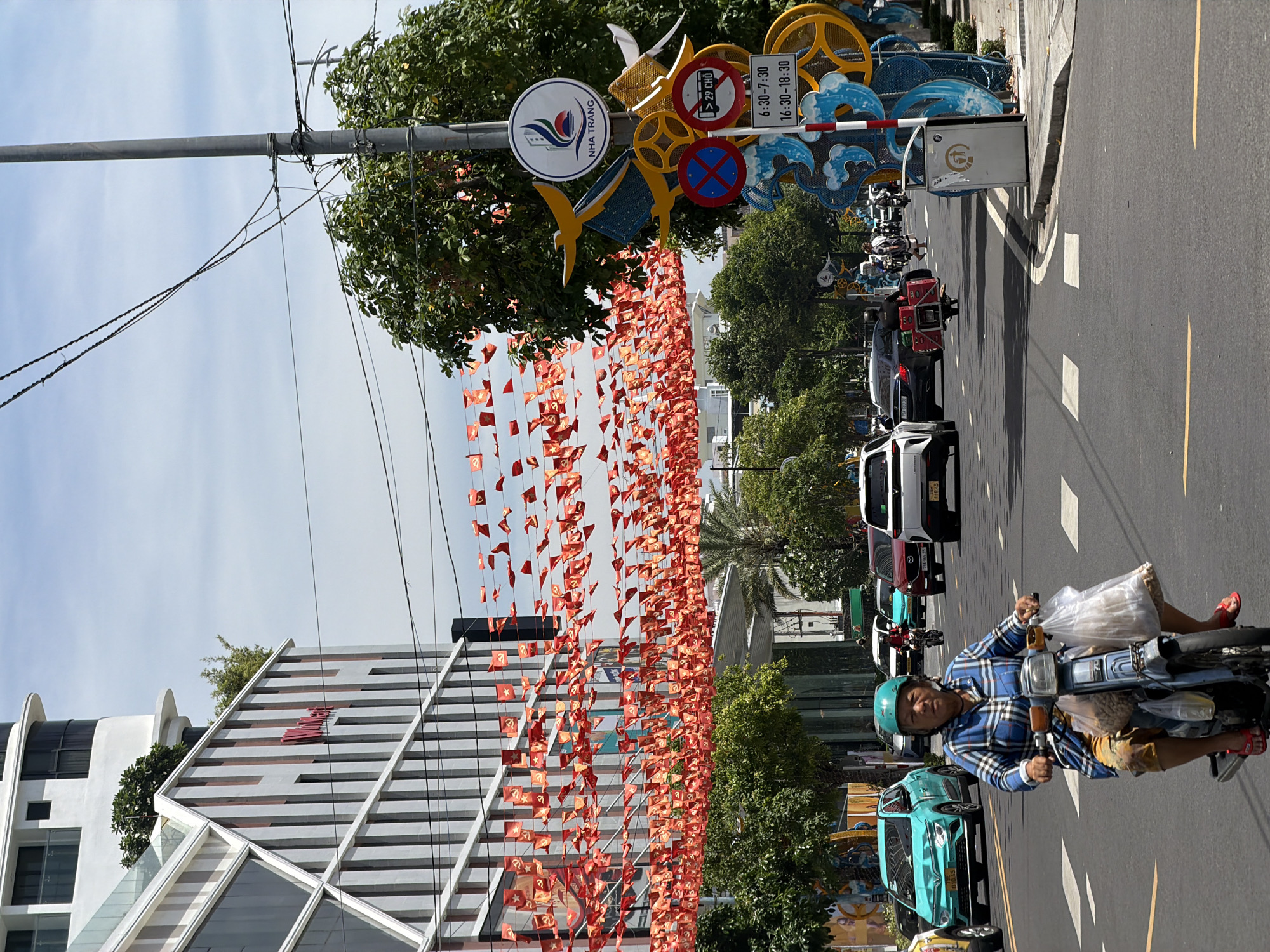 In Nha Trang Ward, Vietnam, a woman wearing a blue and white plaid shirt and a green helmet rides a light blue scooter on a sunny day. She has a string of red flags with yellow stars, likely political banners, overhead. The woman is carrying two plastic bags, one with what appears to be food items. The street is a multi-lane road with other vehicles, including cars and motorcycles. On the right side of the road, a tall pole displays a sign with the Nha Trang city logo and traffic regulations, including restricted hours. Decorative blue and yellow elements, resembling waves and abstract shapes, are mounted on the pole. A modern building with a striking architectural design and glass windows is visible on the left side of the image, with more red flags strung across its facade. The sky is partly cloudy.