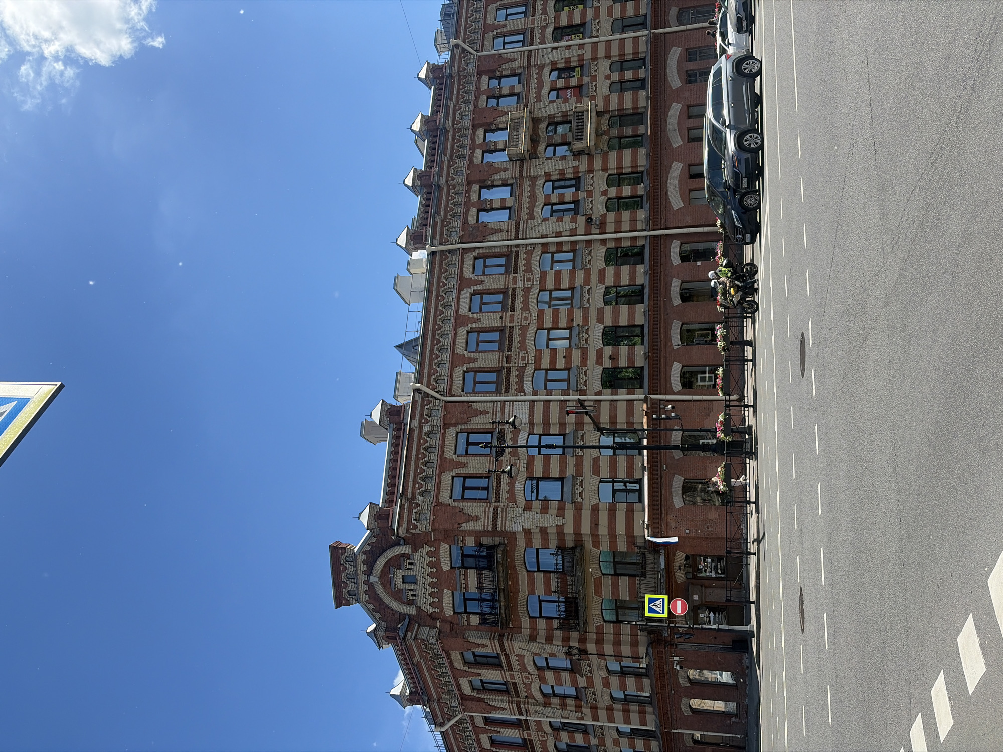 This image shows a street scene in Saint Petersburg, Russia. In the background, a multi-story brick building with white horizontal stripes stands under a clear blue sky. The building features ornate architectural details, including decorative brickwork, arched windows, and balconies. In the foreground, a paved road with lane markings is visible. Several vehicles, including a motorcycle with two riders, a black SUV, a gray SUV, and a white car, are present on the road. A pedestrian crossing sign and a red circular traffic sign are also visible on the sidewalk in front of the building. The overall scene depicts a sunny day in an urban environment.