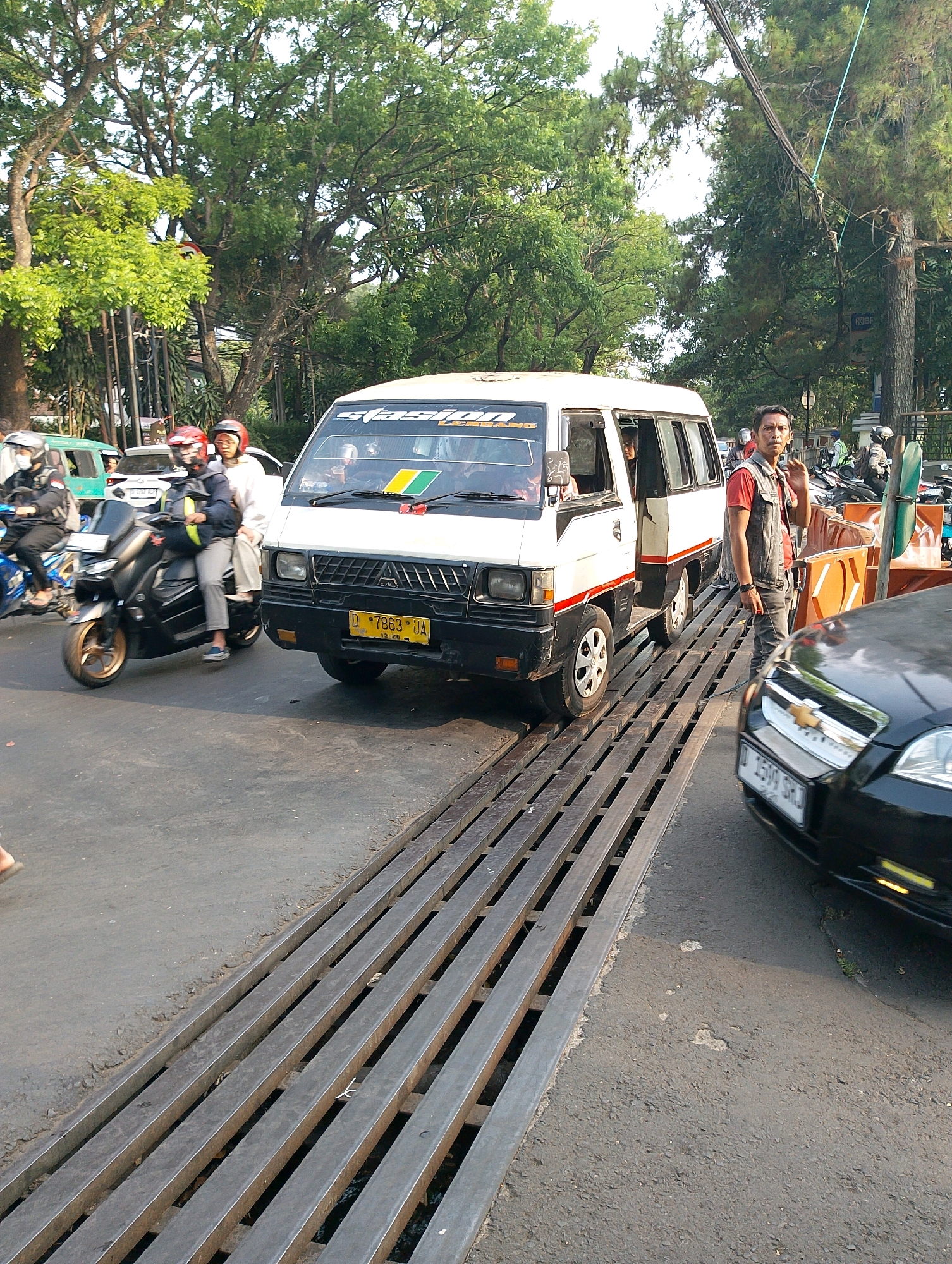 A white minivan with a black and red stripe and yellow license plate number 7863 UA is stopped on a street. A man in a gray vest over a red shirt and dark jeans is standing next to the van, raising his right hand. Behind him, motorcycles are parked. To the left of the van, a black motorcycle with two passengers, one wearing a red helmet and the other a white helmet, is moving forward. A black car is visible in the foreground. The scene appears to be a street in Bandung City, Indonesia, as indicated by the license plate format and typical streetscape.