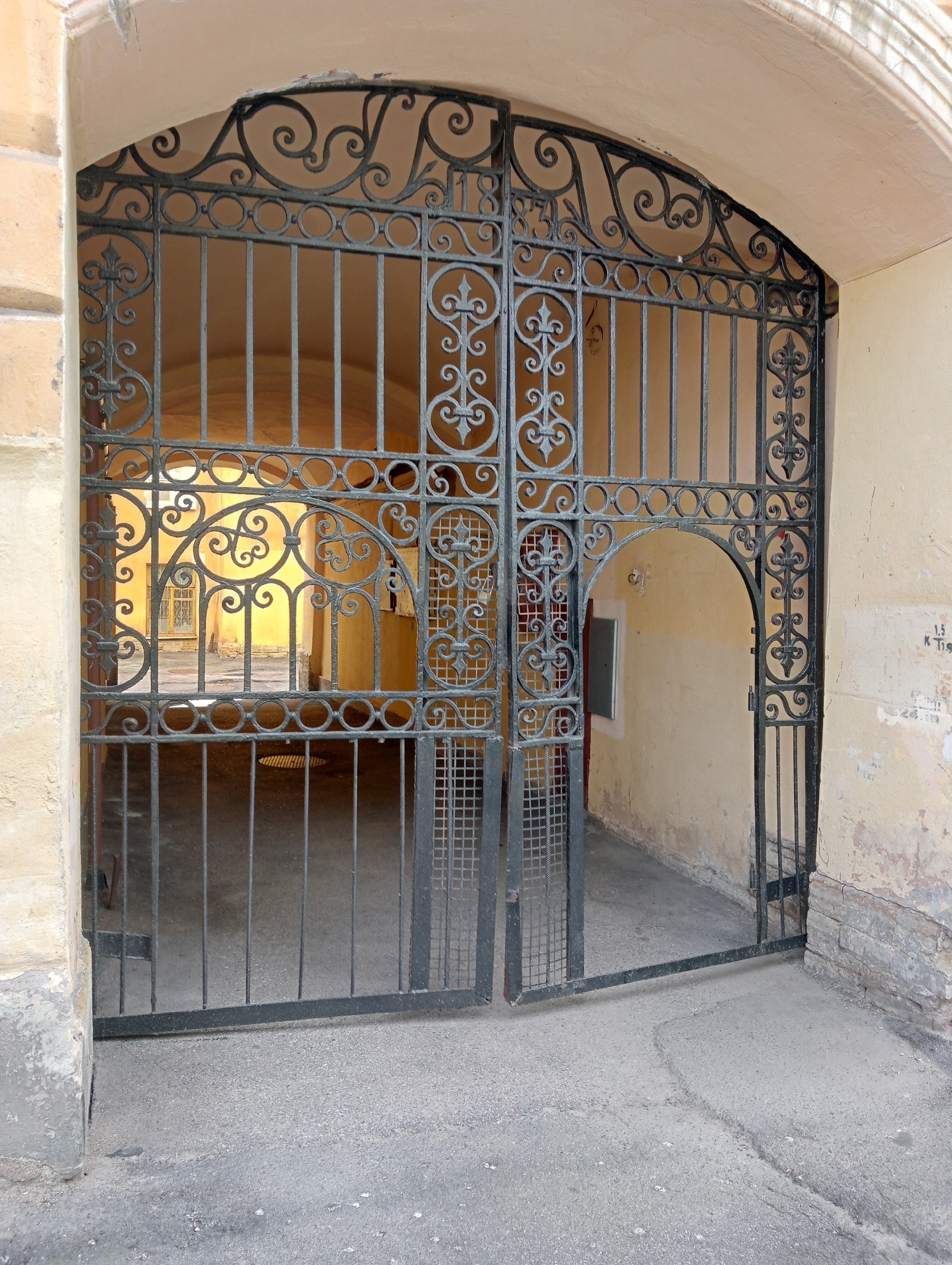 An arched, ornate, black metal gate stands open, leading into a narrow courtyard in Saint Petersburg, Russia. The gate is made of vertical bars and decorative scrolling metalwork. Through the gate, the courtyard is illuminated with yellow light, and a window with a barred grate can be seen in the distance. The walls of the building are a pale yellow, with patches of discolored plaster and brickwork. The ground in front of the gate is asphalt, showing some cracks and debris. Graffiti is visible on the right-hand wall. There are no people in the image.