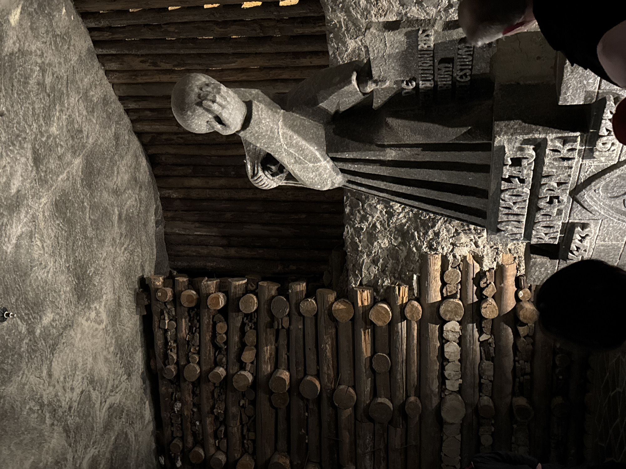 This image shows a sculptural scene in the Wieliczka Salt Mine in Wieliczka, Poland. A gray statue of a person, possibly a miner, stands in a cave-like setting made of salt and wood. The person is facing left with their hand covering their face, apparently looking down. The statue is mounted on a stone with carved lettering in Polish. Behind the statue is a wall of stacked timber. To the left is a large salt formation. People's heads are visible in the upper-right corner.