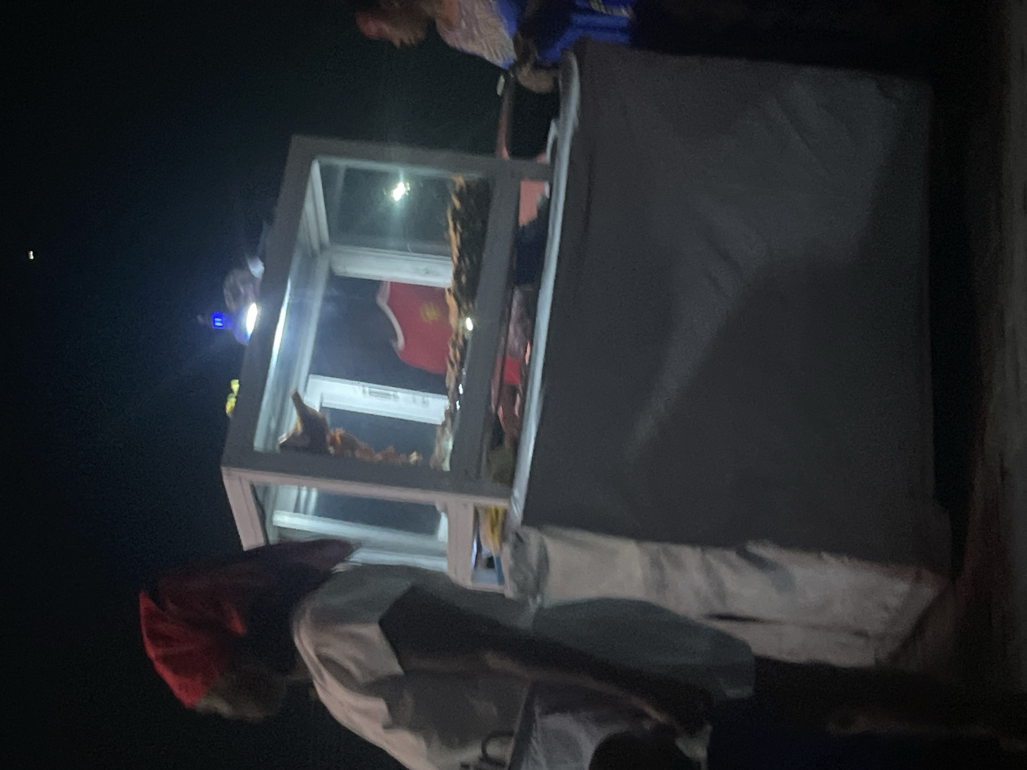 The image shows a food vending cart at night, likely in Lugbe, Nigeria. The cart has a glass display case containing food items. A person wearing a red head covering and a white shirt is visible near the front of the cart. Another person is seen in the background on the right side of the image. The cart is covered with a white cloth. The background is dark.