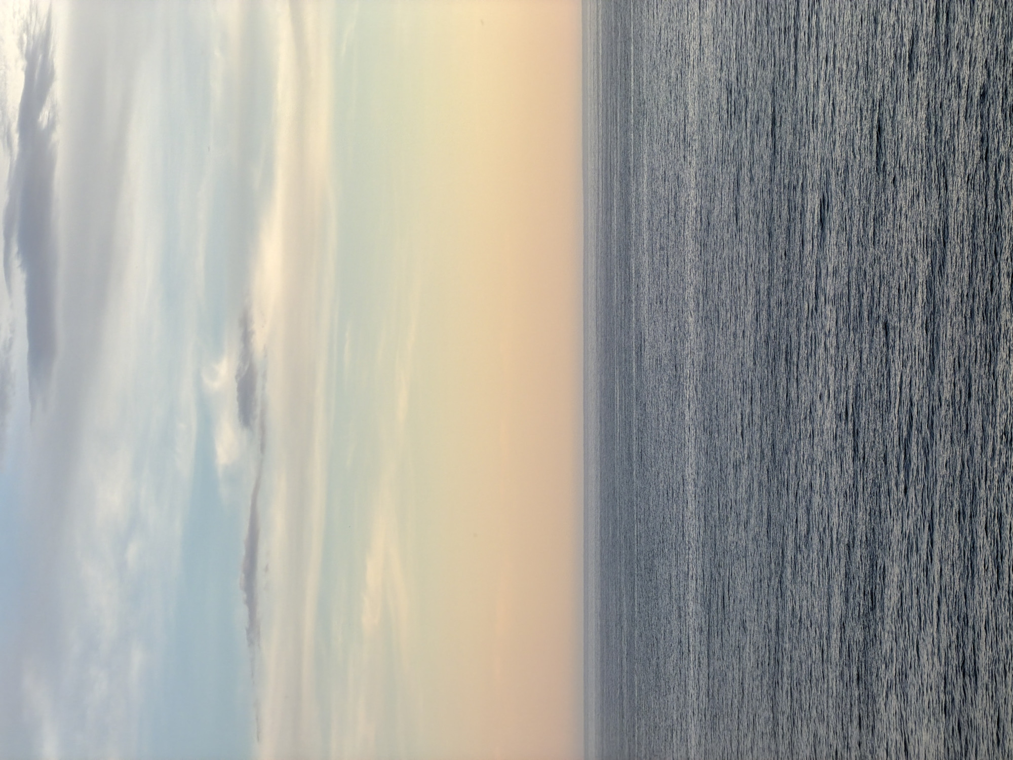 The image shows a view of the sea and sky at Petrovac na Moru, Montenegro. The sky on the left side of the image is light blue with scattered clouds. The sky then transitions to a uniform light yellow color. The sea, which occupies the right half of the image, is gray and rippled.