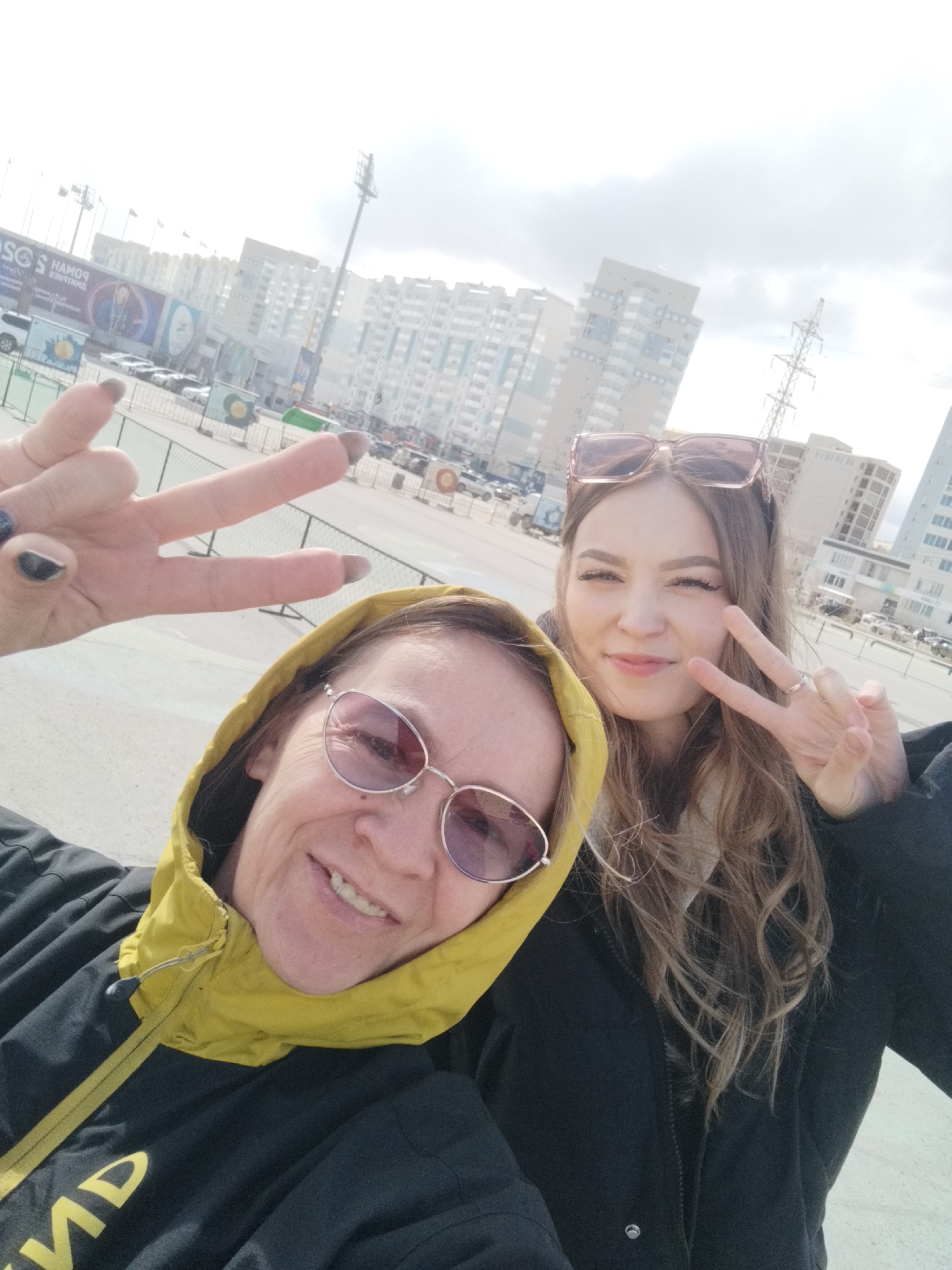 Two women pose for a selfie, both flashing peace signs. The woman on the left is older, wearing a black jacket with a yellow hood and rose-tinted sunglasses. The younger woman is on the right, wearing a black jacket and sunglasses. Both are fair-skinned with light brown hair. Behind them, buildings and cars are visible, as is a power grid tower. The location appears to be outdoors in Yakutsk, Russia.