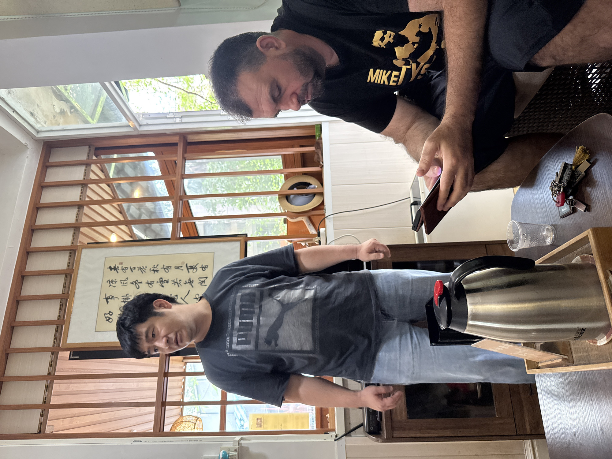 The image depicts two men indoors, possibly in a cafe or lounge area in Huizhou, China, given the presence of Chinese calligraphy on the wall.
The man on the left, wearing a dark grey Puma t-shirt and light blue jeans, stands with his body angled slightly to the right, looking off-camera with a neutral expression. He appears to be in his late 30s or early 40s with short black hair. Behind him, a wooden cabinet with glass doors is visible, containing what appear to be bottles. To his left, a large stainless steel thermos with a black lid and handle sits on a wooden tray, accompanied by a small plastic cup and a few keys.
The man on the right, wearing a black t-shirt with a gold graphic and the word "MIKE" prominently displayed, is seated and looking down at a smartphone he is holding in his hands. He has short brown hair and a beard. His left leg is crossed over his right. The environment features large windows with wooden frames, allowing natural light to enter. The walls are white, with some wooden paneling. The overall atmosphere suggests a relaxed, casual setting.