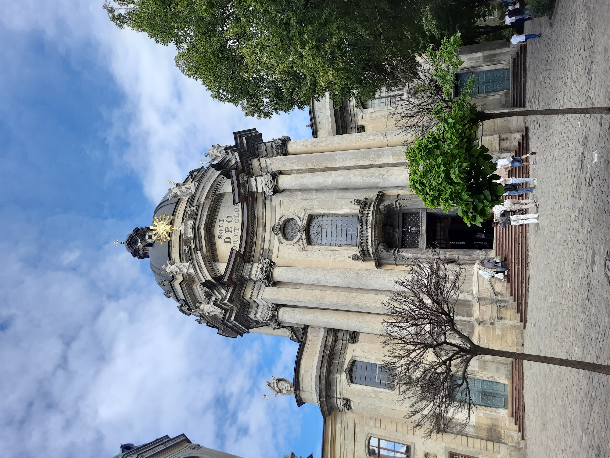 The image captures a frontal view of St. George's Cathedral in Lviv, Ukraine, a prominent Baroque Roman Catholic cathedral. The cathedral features a large, imposing dome topped with a golden cross and a prominent golden starburst. Sculptural figures adorn the facade, and grand columns support the structure. The entrance to the cathedral is reached by a wide set of stone steps, with people visible ascending and descending. In the foreground, a paved courtyard is visible, with two trees in the mid-ground. One tree is bare and has a spindly trunk and branches, while the other is full of lush green leaves. Several individuals are seen walking in the courtyard and near the entrance, some appearing to be tourists taking photos. The sky above is partly cloudy, with a mix of blue sky and white clouds. Lush green trees are visible on the right side of the image, framing the cathedral. The overall scene suggests a daytime visit to a significant historical and religious landmark.