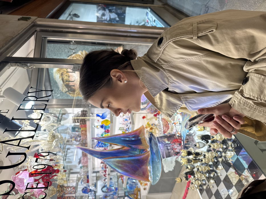A young adult female, shown in right profile, stands outdoors, intently observing the display in a shop window. She has medium-dark brown hair tied back in a bun and wears a light beige trench coat with its collar elevated. A small, circular stud earring is visible in her right ear. Her hands are clasped, holding a smartphone with a transparent case at waist level; a plain silver ring adorns the ring finger of her right hand.
The shop window, labeled "GALLERY" and "SINCE 1979" on the glass, showcases numerous Murano-style glass art pieces. Prominently featured is a large, iridescent blue and purple glass sculpture resembling a sailboat with two sails, positioned in the mid-ground. In the lower foreground, a glass chessboard with clear and dark-colored pieces is displayed on a reflective surface. Behind these, shelves hold various smaller colorful glass figurines, vases, and decorative items. Chandeliers made of glass are visible hanging within the shop, reflected and seen through the upper part of the window. Reflections of architectural elements and a partial image of a classical artwork are visible on the shop window's surface. The setting is a narrow pedestrian passage in Venice, Italy, consistent with a shop specializing in Murano glass.