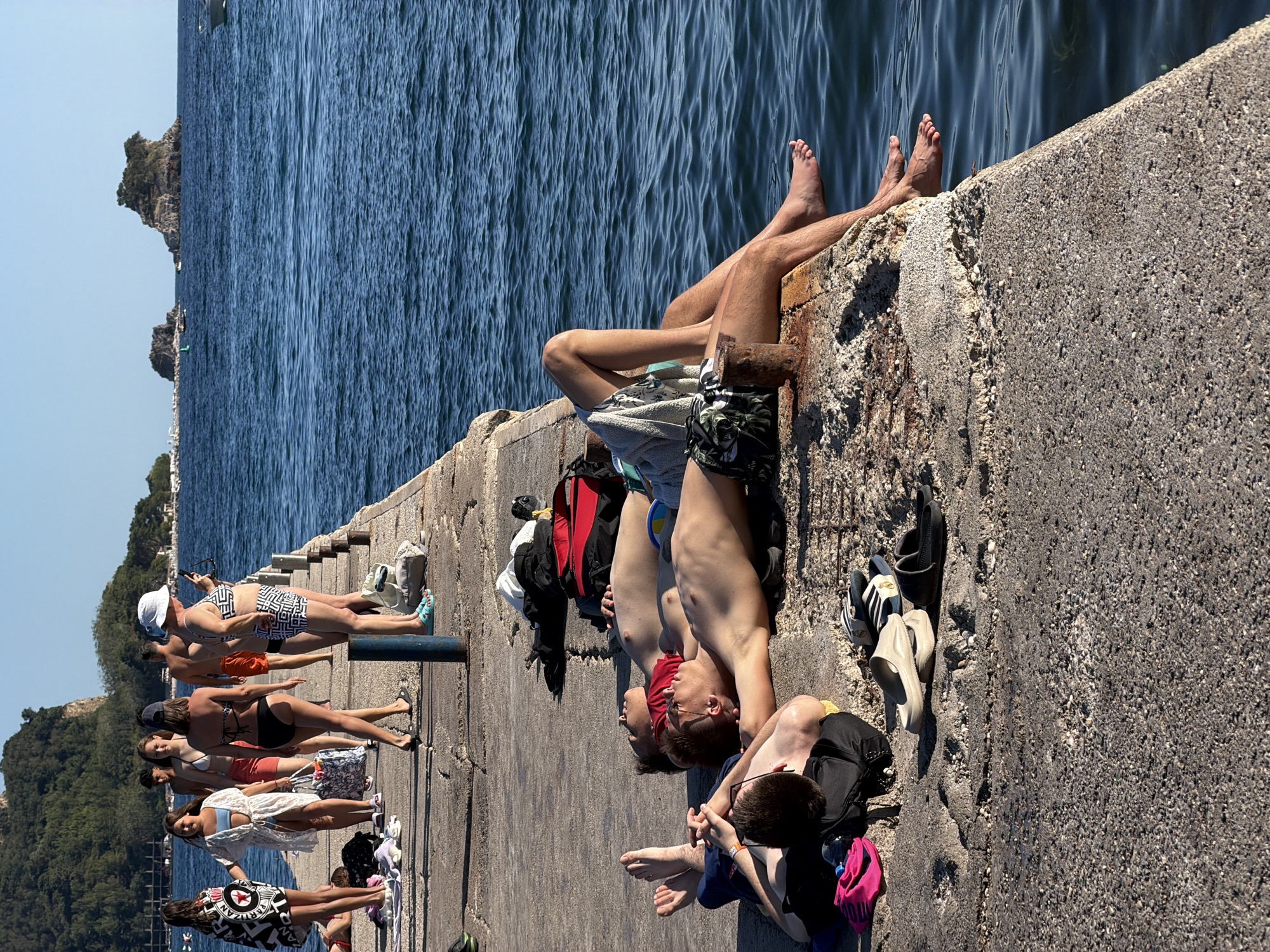 In Budva, Montenegro, on a sunny day, a concrete pier extends into the blue Adriatic Sea. Several individuals are enjoying the warm weather. In the foreground, three young men are lying on the pier, some sunbathing and others resting. Bags and sandals are scattered around them. Further down the pier, a group of people, including men and women, are walking and standing, some wearing swimwear. One woman in the distance is taking a photo with her phone. The background shows a green, rocky hillside and small islands in the sea. Boats are visible on the water. The overall scene depicts a relaxed atmosphere at a popular seaside location.