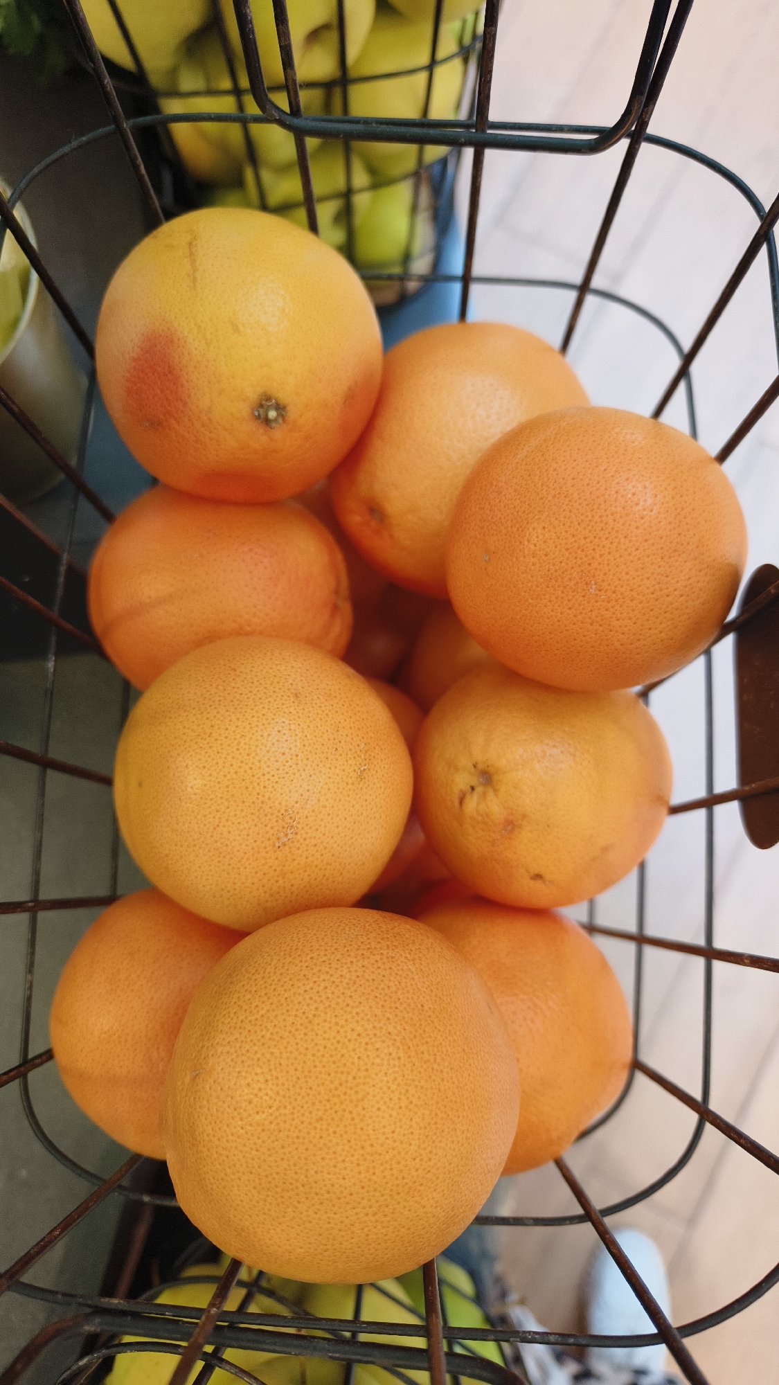 A close-up, top-down view shows a wire basket filled with grapefruits. The grapefruits are predominantly orange with some exhibiting a reddish blush. They are arranged in multiple layers within the basket. The basket itself appears to be made of dark-colored wire with a slightly rustic aesthetic. In the background, out of focus, are some green fruits, possibly apples or limes, also contained within a basket. The surface beneath the basket appears to be a light-colored, possibly tiled floor. The lighting is bright, casting minimal shadows. The scene is devoid of any people or discernible human activity. This image was taken at the Vösendorf Shopping City Süd in Vösendorf, Austria.