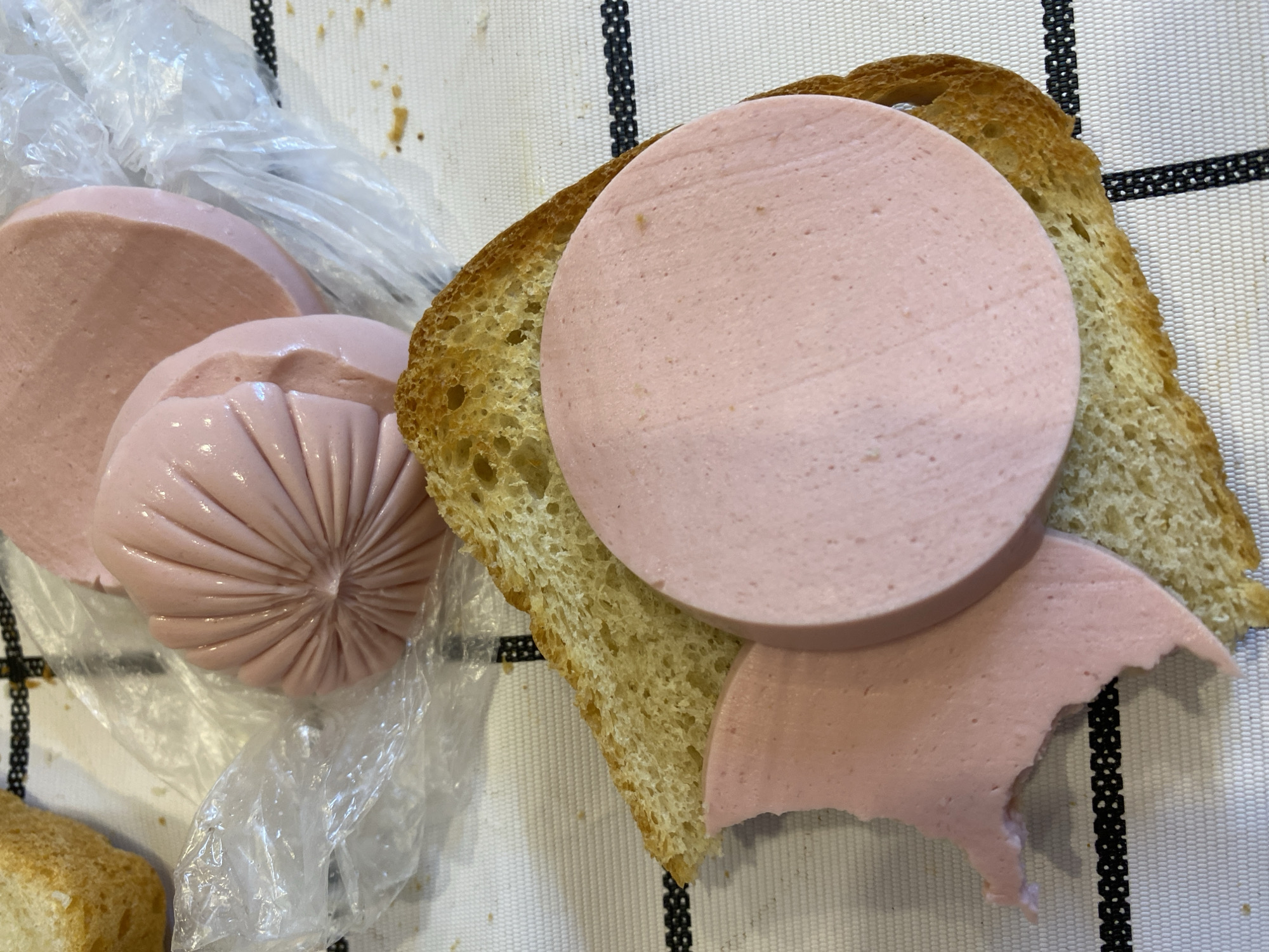 The image shows a close-up, high-angle shot of a sandwich being prepared on a white, textured tablecloth with a black striped border. The sandwich consists of a slice of toasted bread topped with several thick slices of pink bologna sausage. Some of the bologna appears to have been bitten or torn. In the background, more bologna sausage is visible, still partially in its plastic packaging, along with another slice of bread. The scene is well-lit, with no people visible. The location is Lomonosov, Russia, as indicated by the context provided.