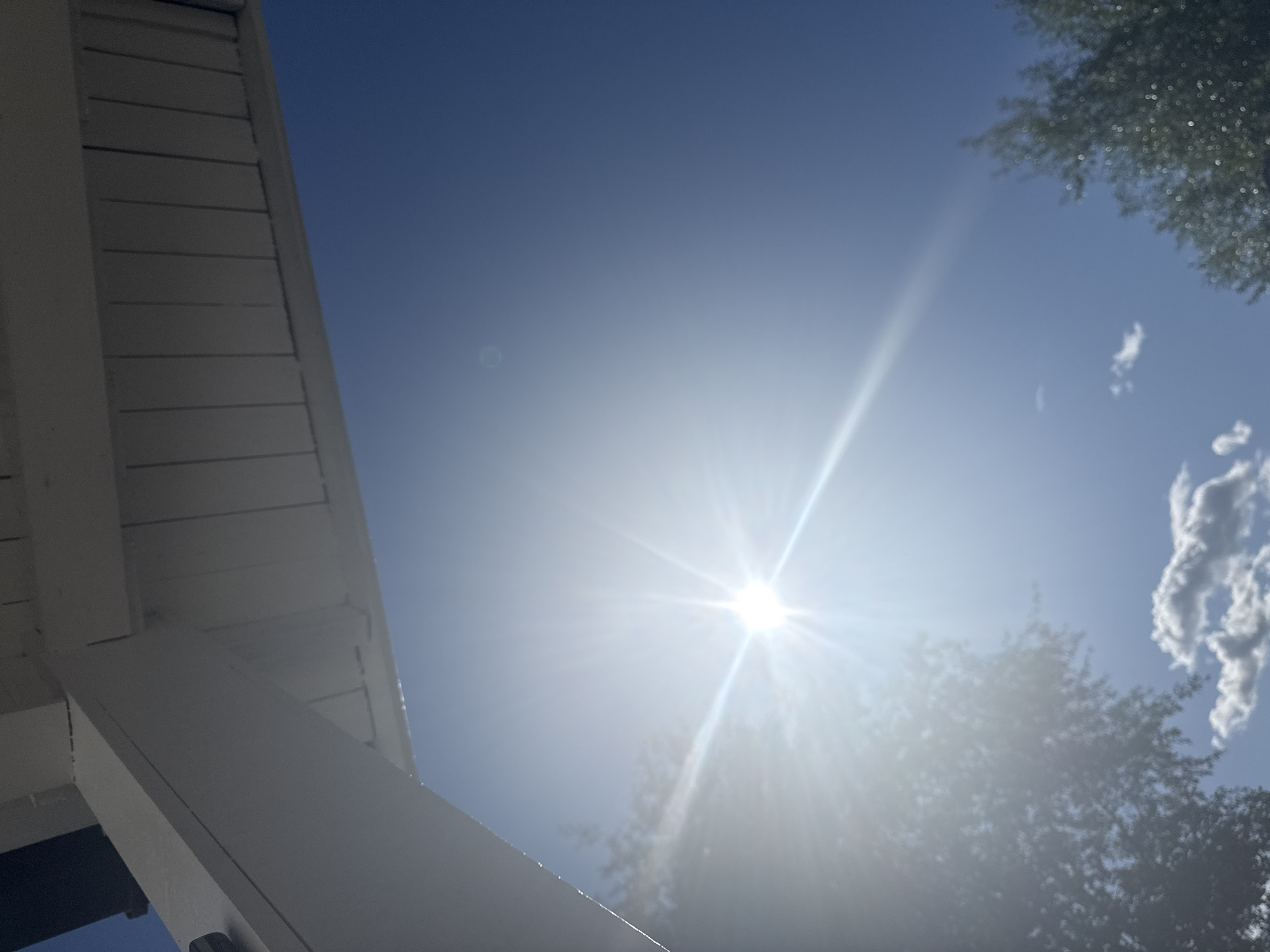 This is a low-angle, wide shot of a bright sunny sky taken from under a white wooden overhang, possibly a porch or awning. The sun is prominently featured in the center-left of the frame, emitting bright rays and lens flare. The sky is a clear, deep blue, with a few wispy clouds visible lower down. The edge of the wooden structure, with its slatted ceiling and white support beams, occupies the top portion of the image. There are hints of trees with green foliage at the bottom left and right edges of the frame, suggesting an outdoor setting. There are no people or specific landmarks visible to pinpoint a location within Moelv, Norway.