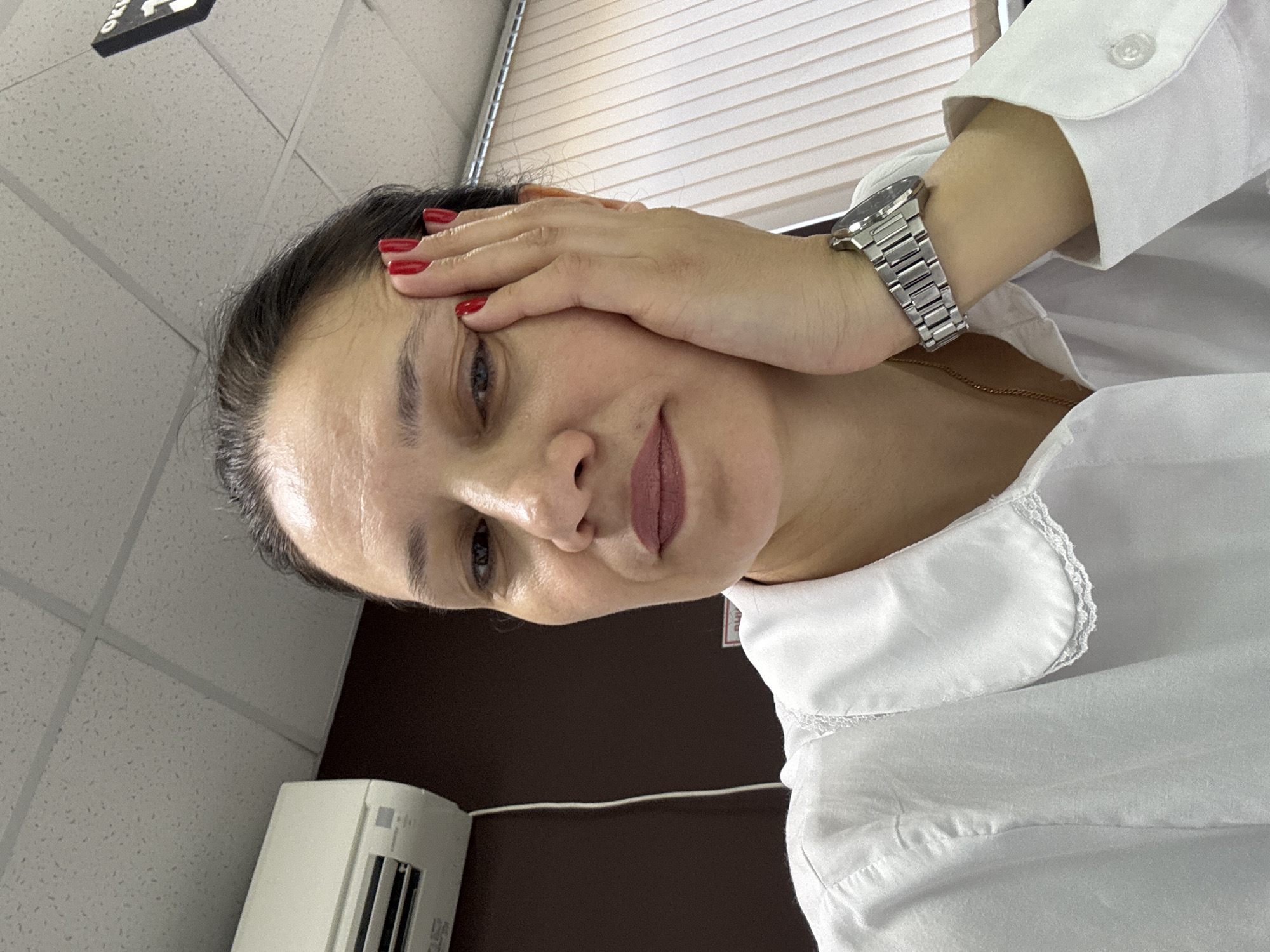 A woman with dark hair and red nail polish rests her hand on her forehead. She wears a white button-down shirt and a silver wristwatch. The background includes a window with blinds, a tiled ceiling, and an air conditioning unit. The location is Chaikovsky, Russia.