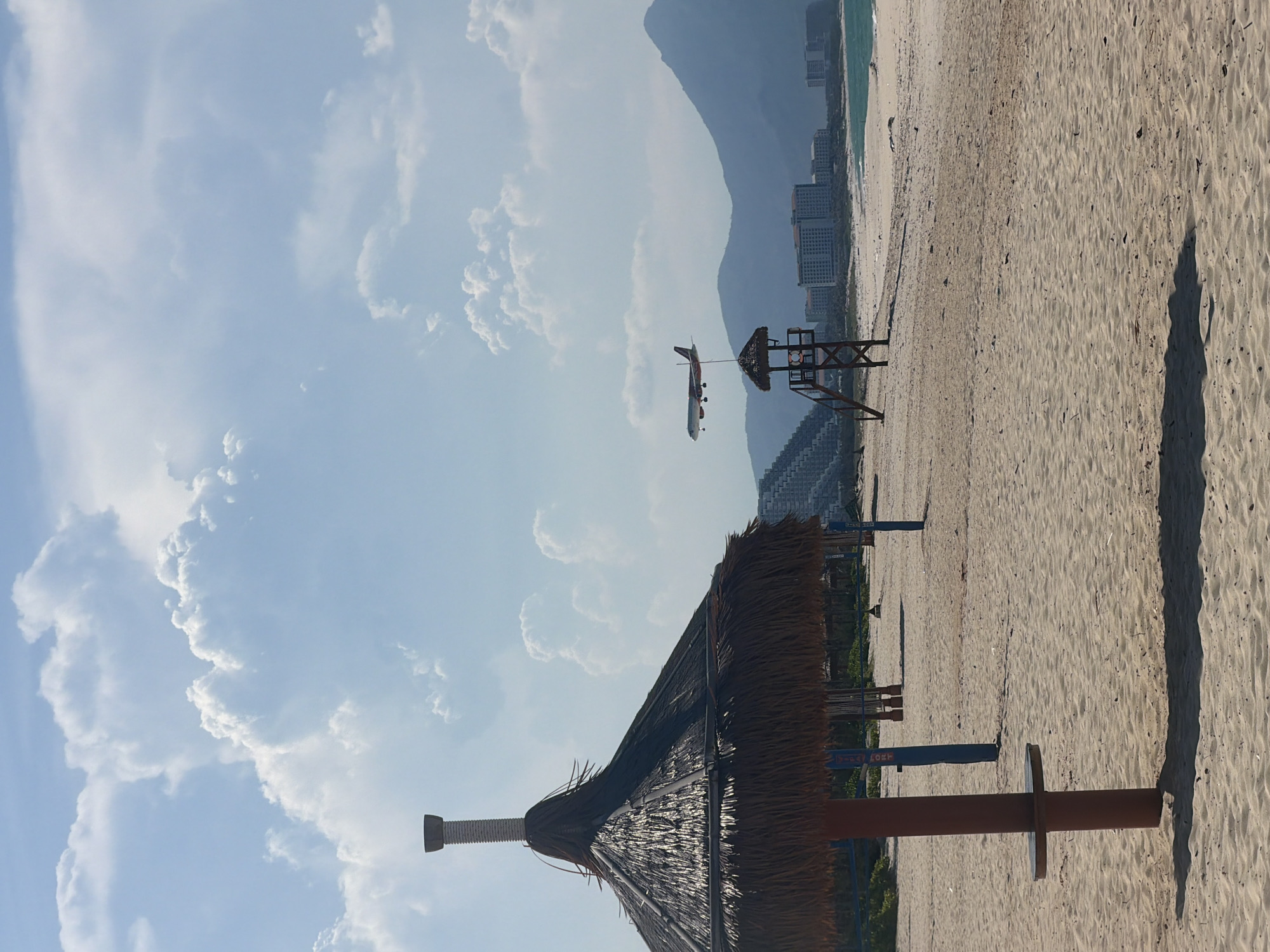 A photograph captures a beach scene in Bac Cam Ranh Ward, Vietnam, with an airplane in flight. In the foreground, a thatched umbrella stands on a sandy beach, casting a shadow. A wooden lifeguard stand is visible further down the beach, with buildings and mountains in the background. The sky is partly cloudy.