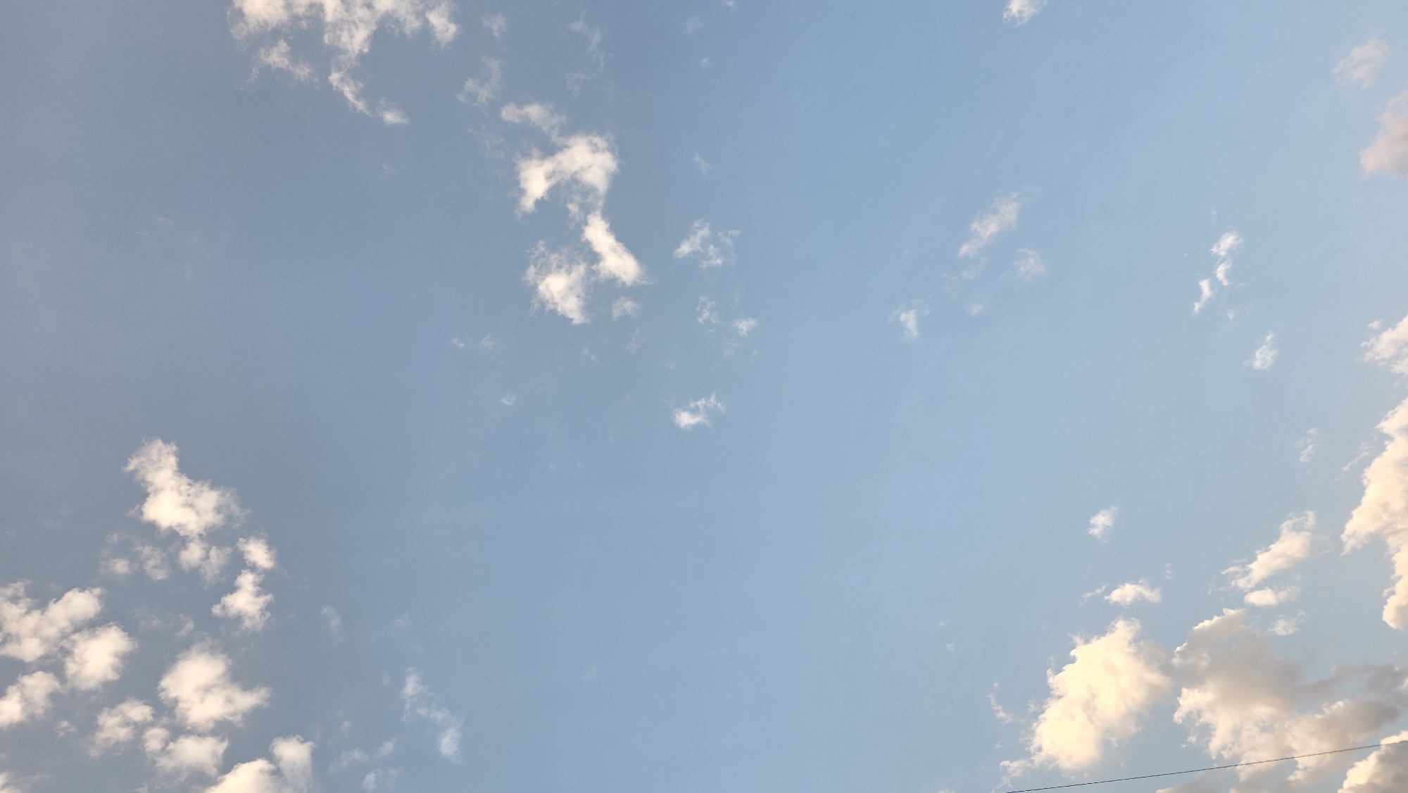 The image shows a clear blue sky with scattered white clouds. There is a thin wire visible in the lower left corner of the frame. The image appears to be taken in an outdoor environment. No people, subjects, or actions are discernible.