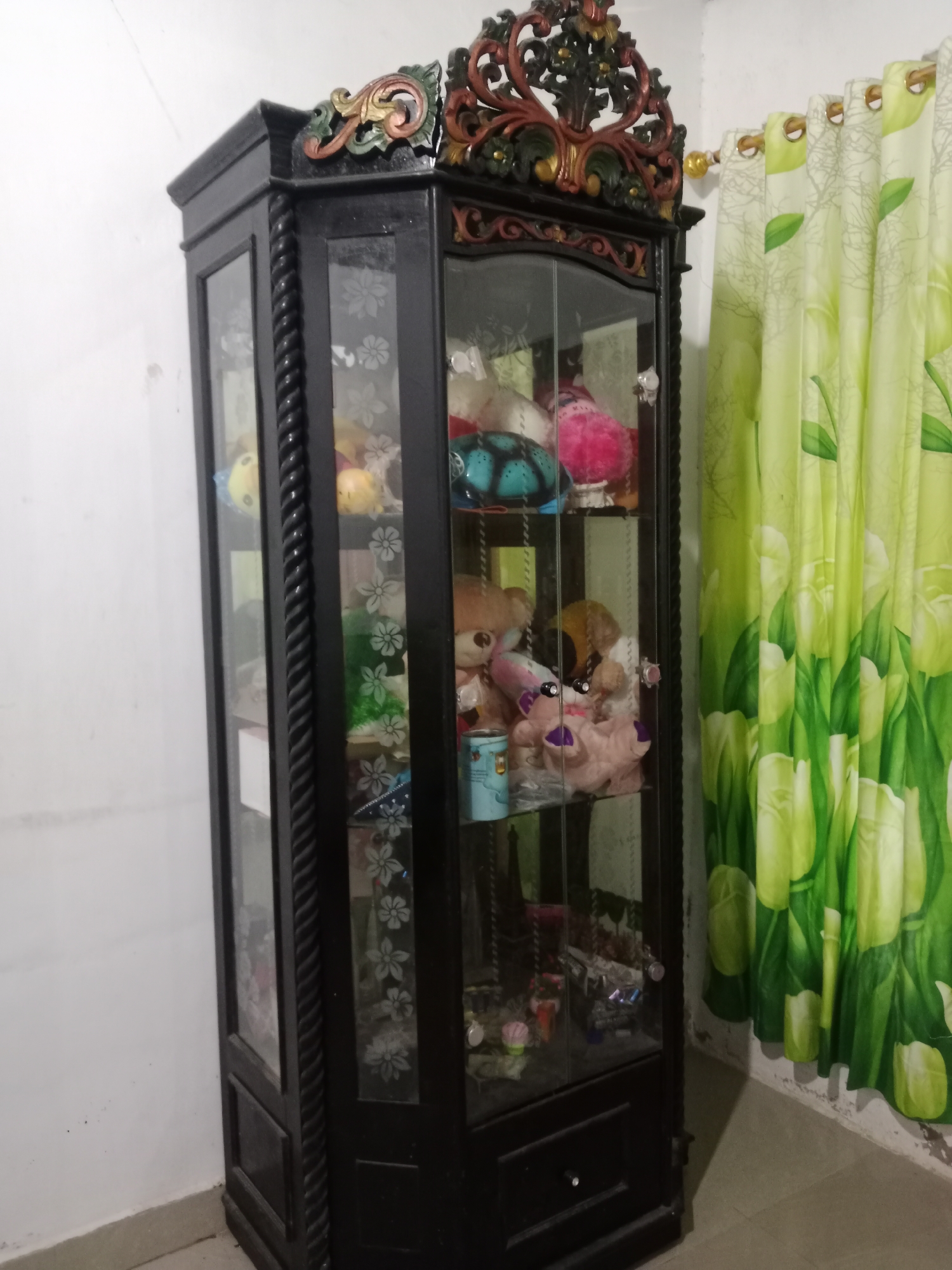 An ornate, dark wood curio cabinet stands against a white wall in Tambakcekur, Indonesia. The cabinet has glass doors with etched floral patterns, revealing shelves filled with various stuffed animals and small decorative items. The top of the cabinet features elaborate carved wood ornamentation in red and gold. To the right of the cabinet, bright green curtains with a floral leaf pattern are drawn. The floor appears to be tiled. The scene suggests a domestic interior setting.