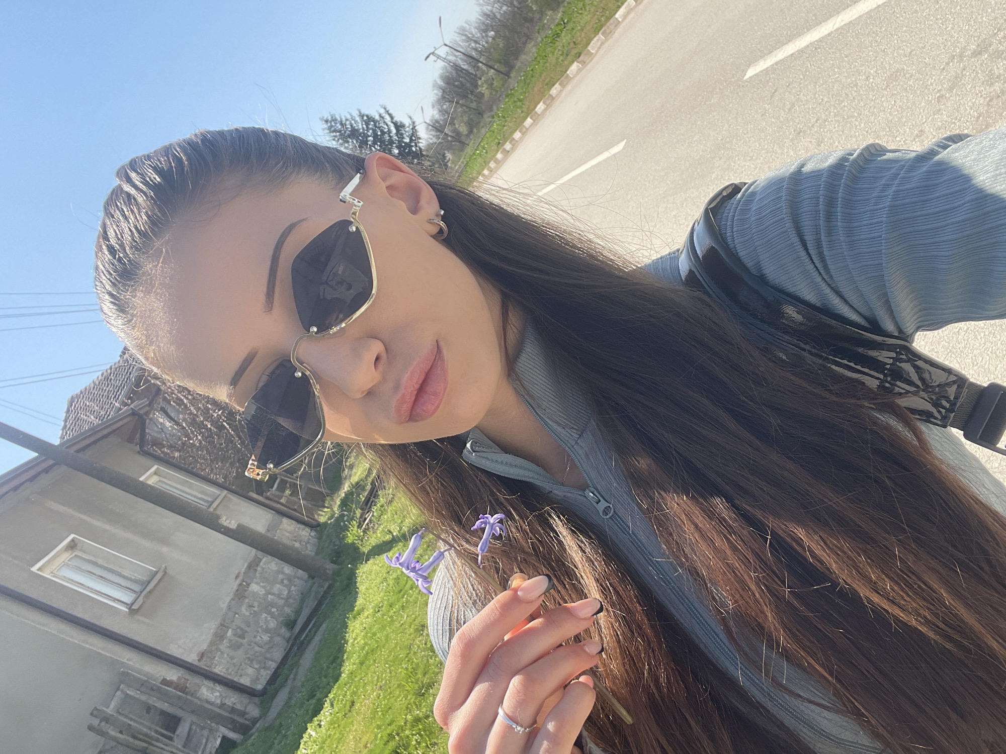 A medium shot shows a young woman holding a small, purple flower. She has long, dark brown hair, fair skin, and is wearing sunglasses and a gray zip-up top. The background shows a street in Somovit, Bulgaria, with a two-story building with gray walls and windows and a rural road with green grass beside it.