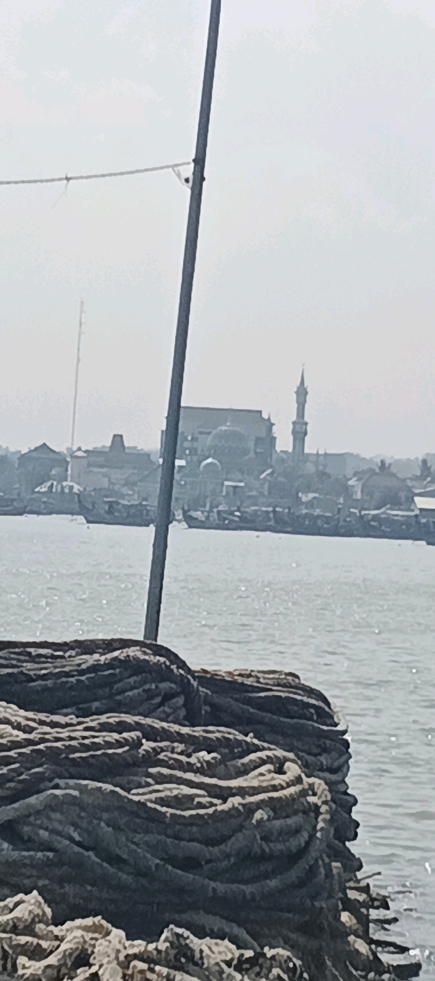 The image depicts a hazy daytime scene at the Blimbing, Indonesia waterfront. In the foreground, a large coil of thick, dark rope is positioned on the right, extending into the lower portion of the frame. The rope is densely packed and textured. A tall, slender pole, likely part of a boat or structure, runs vertically through the center of the image. In the middle ground, a body of water is visible, shimmering with sunlight. Beyond the water, a city skyline is discernible through the haze. Prominent structures include buildings with domes, a minaret, and what appears to be a tall antenna or tower in the distance to the left. Several boats are scattered on the water and docked along the shore. The overall atmosphere is bright but obscured by atmospheric conditions.