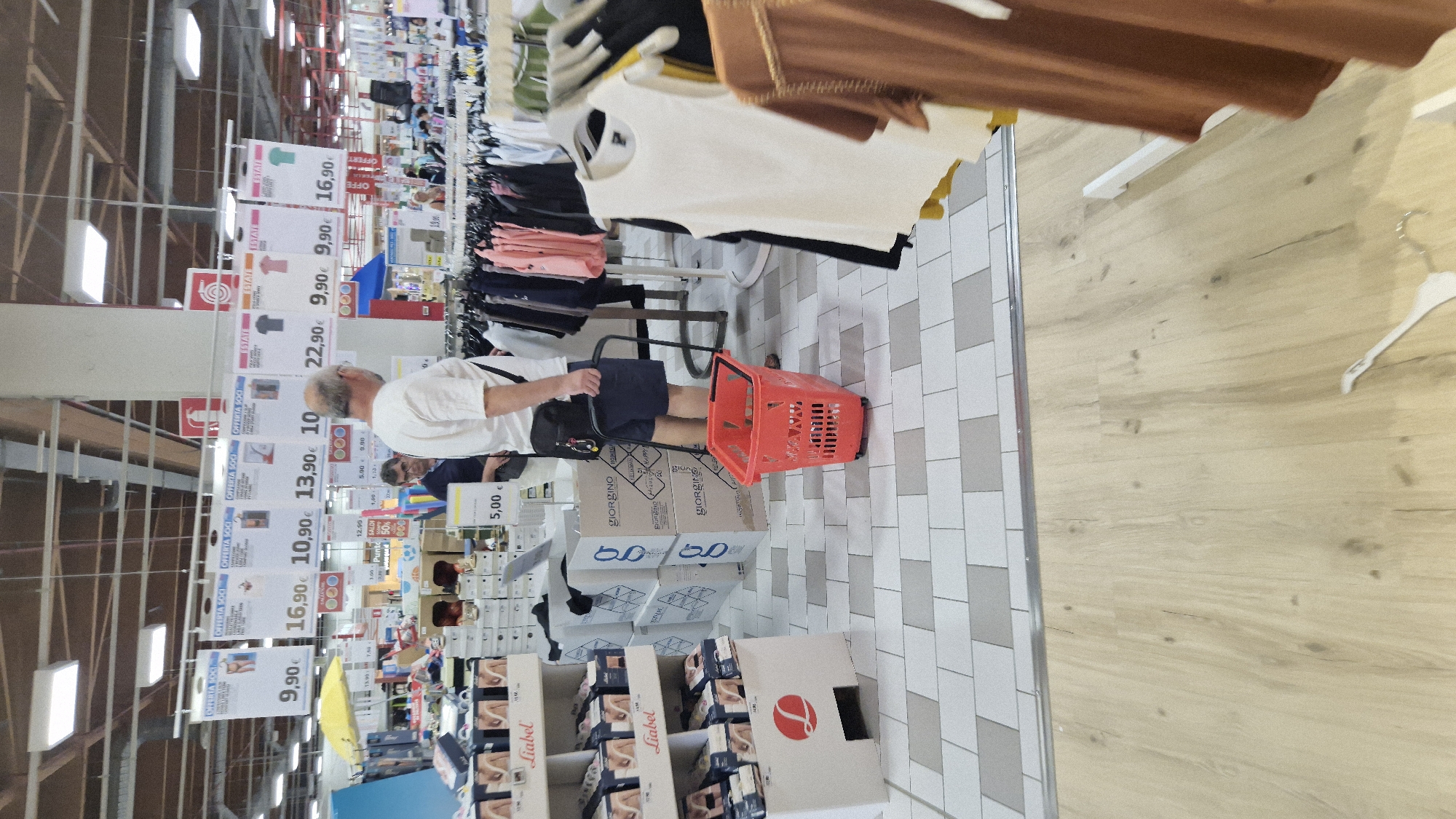 In Taranto, Italy, within a well-lit supermarket with high ceilings and exposed ductwork, a man wearing a white t-shirt and dark shorts is standing near a red shopping basket. He has a black fanny pack around his waist. The floor is a tiled pattern of white and grey squares, transitioning to wood laminate. In the background, clothing racks are visible, displaying various garments. Several price tags are suspended from the ceiling, advertising items with prices like 9.90€, 16.90€, and 10.90€. Boxes are stacked on the floor, some with the "Liabel" and "Giorgino" brand names visible. Another man, wearing a blue shirt, is partially visible behind the man with the fanny pack. The scene suggests a typical shopping environment in a retail store.