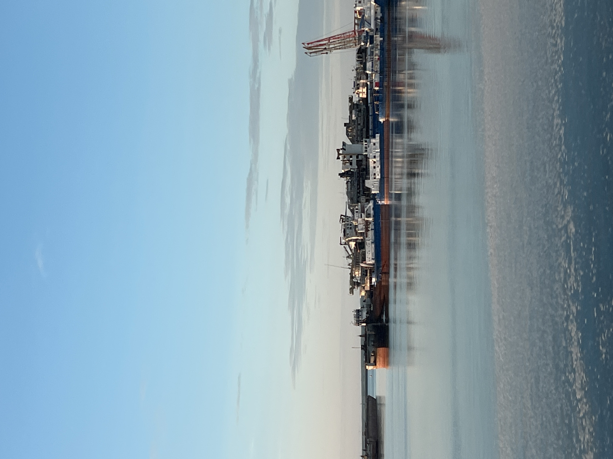 The image displays an exterior daytime scene of a cluster of industrial vessels situated on a calm body of water in Chaikovsky, Russia. The upper approximately 55% of the frame features a light blue sky with scattered, thin, stratiform clouds, predominantly located in the middle distance. A low, indistinct landmass forms a subtle horizon line. The lower approximately 45% of the frame is occupied by the placid water, which exhibits reflective properties, mirroring the sky and the silhouettes of the vessels. The foreground water surface shows a subtle textured pattern of small ripples.
A dense group of approximately ten to fifteen ships and barges occupies the middle ground. These include:
* A dark-hulled vessel partially visible on the far left, extending towards the frame's center.
* Multiple cargo vessels or river tankers moored together, featuring hulls in varying shades of dark brown, orange, and blue, topped with white or light-colored superstructures.
* One prominent vessel in the mid-center has a distinct white pilothouse structure.
* On the far right, a large, multi-segment industrial crane, predominantly red, rises vertically, appearing to be part of a vessel or a shore-based facility.
The vessels exhibit characteristics consistent with working craft, including visible decks, cabins, and rigging. No human figures are identifiable on board or within the surrounding environment. The lighting suggests either early morning or late afternoon, characterized by a soft, slightly warm illumination on the vessels and partial under-lighting of some clouds. The overall scene indicates an industrial maritime or riverine setting, likely a port, shipyard, or vessel lay-up area within the specified city.