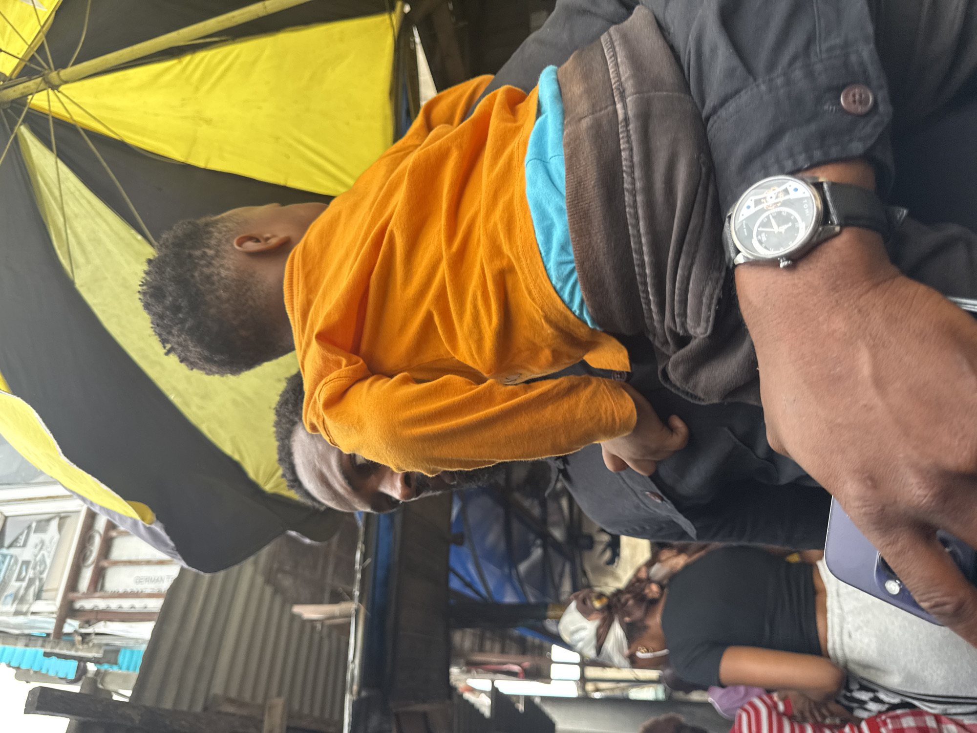 In Lekki, Nigeria, a man carries a young child on his shoulders. The man wears a black jacket over a black shirt, and a wristwatch on his left wrist. The child is dressed in an orange long-sleeved shirt and blue underwear, with grey pants visible around the waist. A black and yellow umbrella is held above them, providing shade. In the background, a woman with a white headscarf and a black t-shirt is seen, along with other individuals and structures that suggest a bustling urban environment. The scene appears to be captured during daylight hours.