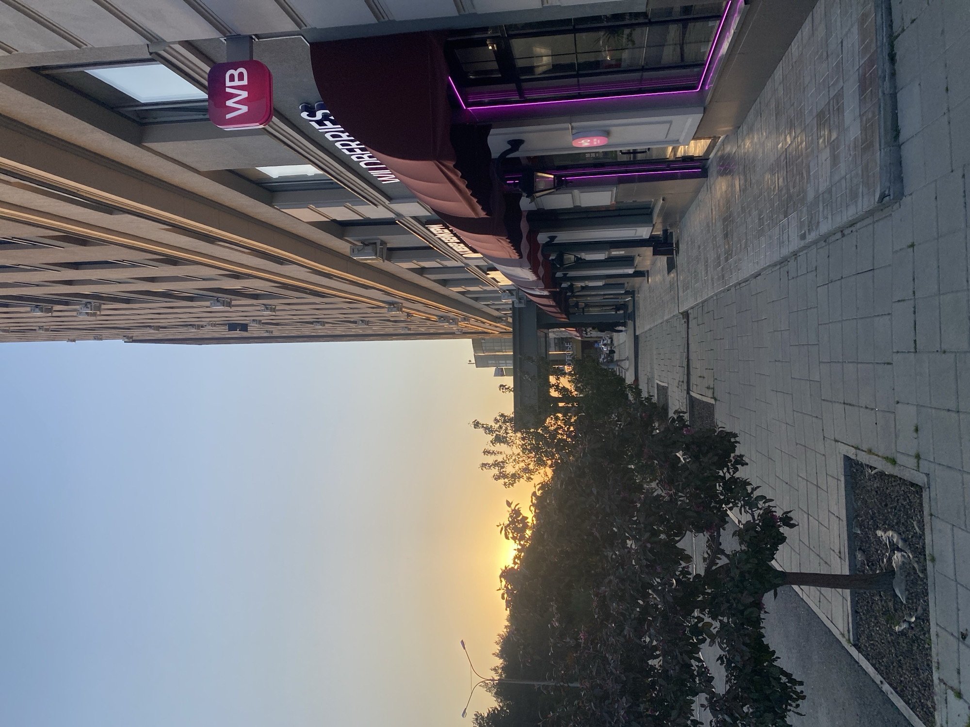 The image shows an urban scene in Yekaterinburg, Russia at sunset. The building on the left has a sign with "WB" on it. The building has a dark red awning with "VAN CREPES" printed on it. There are lights on the right side of the awning. There is a sidewalk on the right side of the building. A tree is visible in the lower right of the image. The sky is a gradient of light blue to orange with the sun visible through the trees in the center.