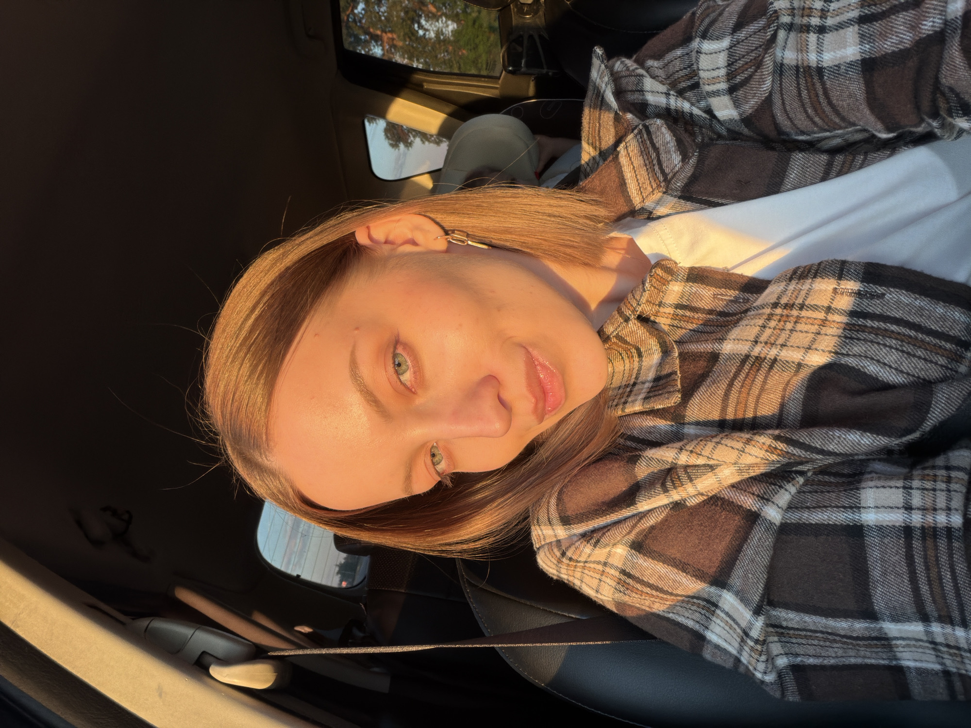 A young woman is photographed from a low angle while sitting inside a car. She is fair-skinned with light brown hair and green eyes. She is wearing a white t-shirt underneath a brown and tan plaid shirt. Golden sunlight streams into the car, illuminating her face and clothing. The interior of the car is visible, including the black leather seats and a window showing foliage outside, suggesting a natural or suburban environment. There is no discernible information about the location being in the city of Rogachovo, Russia.