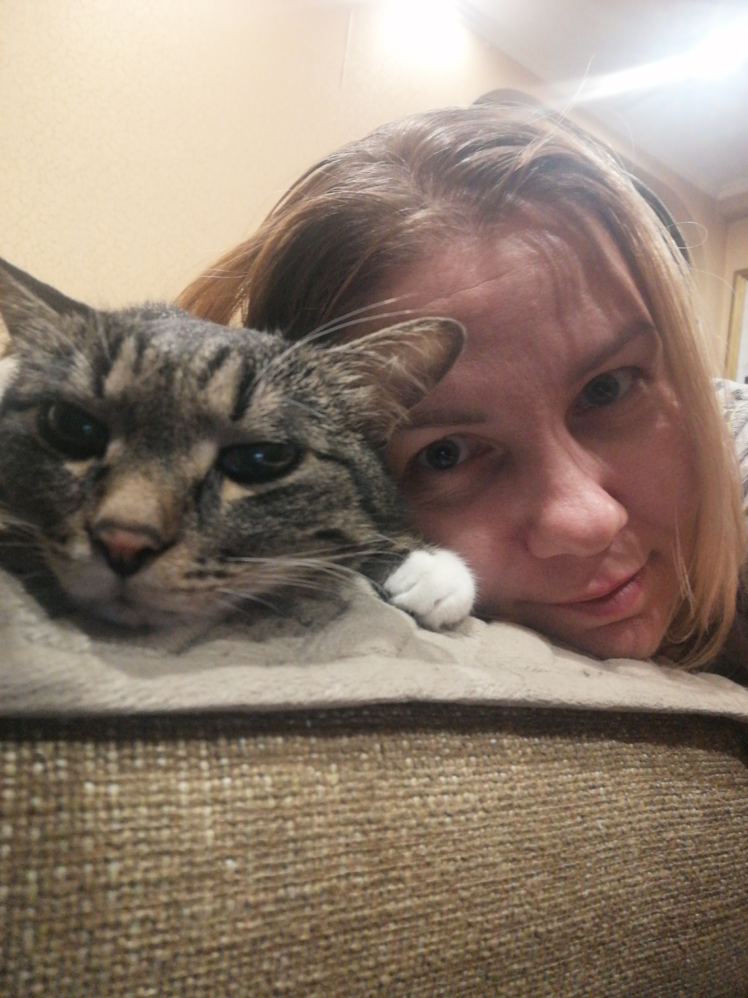 The image is a close-up selfie of a woman and a tabby cat lying down, with the cat's head resting on a textured surface in the foreground and the woman's head behind it. The woman has blonde hair and is looking at the camera with a slight smile. The cat has grey and black stripes and its body is mostly obscured by the woman. The background is a plain, light-colored wall. The scene appears to be indoors, likely in a residential setting. There are no discernible landmarks or context to indicate a specific location in Angarsk, Russia.