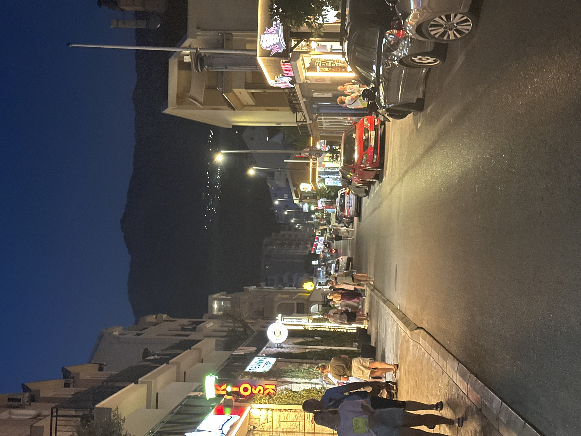 The image depicts a street scene at dusk in Budva, Montenegro, likely along the coast given the presence of mountainous terrain in the background and the general atmosphere of a tourist-oriented town. The street is lined with multi-story buildings housing shops and restaurants, many with illuminated signs. A prominent neon sign reading "KIOSK" is visible on the left side of the street. Several people are walking on the sidewalk, some appearing to be tourists. Cars are parked along the right side of the street and some are driving on the road. The sky is a deep blue, transitioning from twilight. The overall impression is of a lively evening street in a coastal town, with activity and commercial establishments contributing to the ambiance.