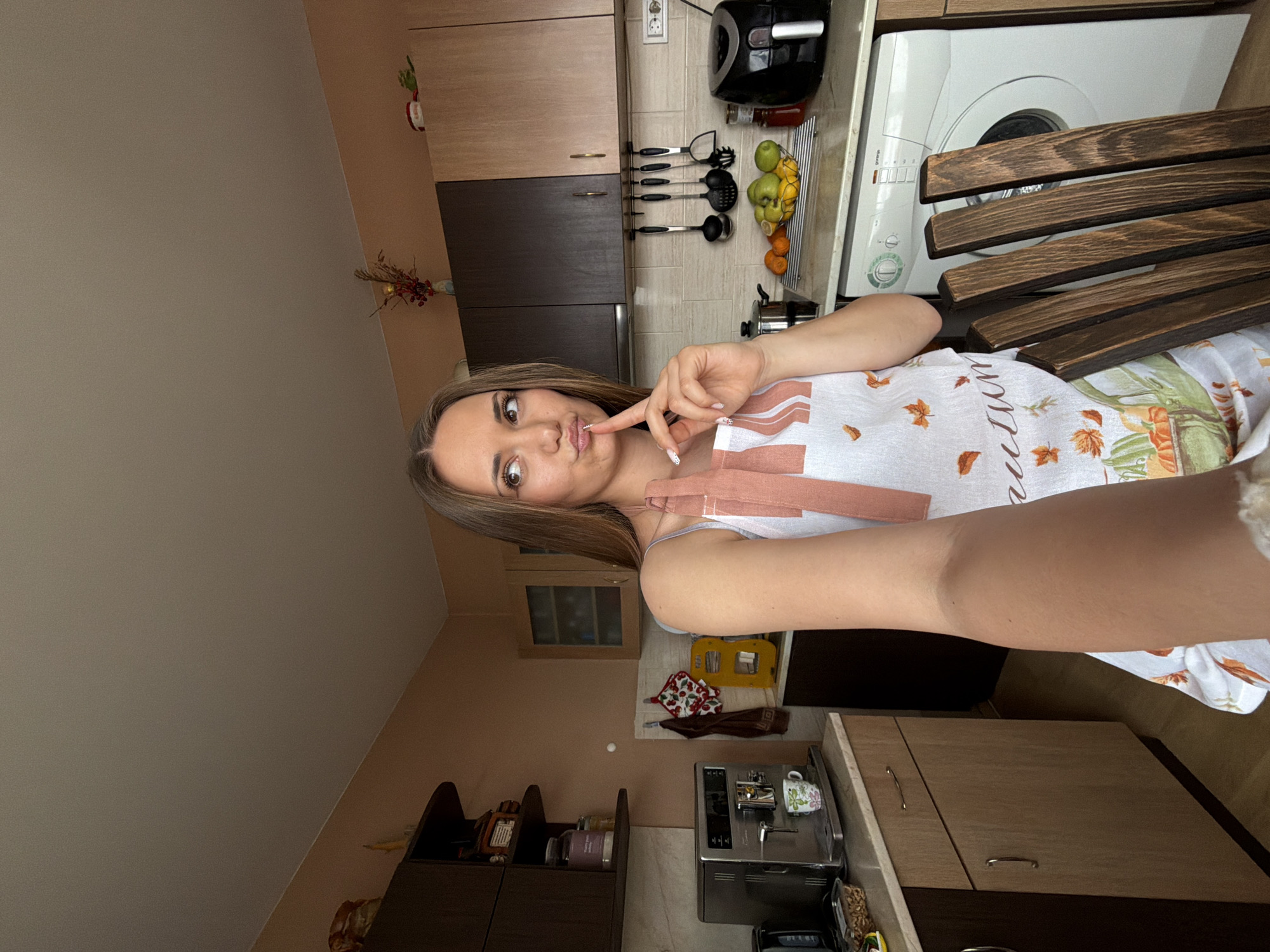 This media file captures a young woman taking a selfie in a home kitchen. The woman, who appears to be in her 20s or early 30s, has long brown hair and is wearing a light-colored top under a white apron patterned with autumn leaves and pumpkins. She is puckering her lips slightly in a "duck face" pose, with her right index finger touching her chin/mouth, conveying a playful or casual mood.
The setting is a well-equipped kitchen. In the background, there are light and dark wood kitchen cabinets. On the counter to her left, kitchen utensils are neatly hung, and a fruit bowl holds green apples and oranges. Further back, an air fryer and a washing machine are visible. The washing machine has the brand "Gorenje" faintly visible on its front panel. To her right, a silver coffee machine with a patterned mug sits on the counter, next to what appears to be a spice rack or small shelves holding jars. A small plant or decoration with red berries adds a touch of color near the cabinets.
On the apron, a cursive word, likely "autumn," is partially visible. The lighting suggests it is daytime. The general appearance of the kitchen, including the style of outlets, is consistent with a European setting, aligning with the provided context of Buhovo, Bulgaria.