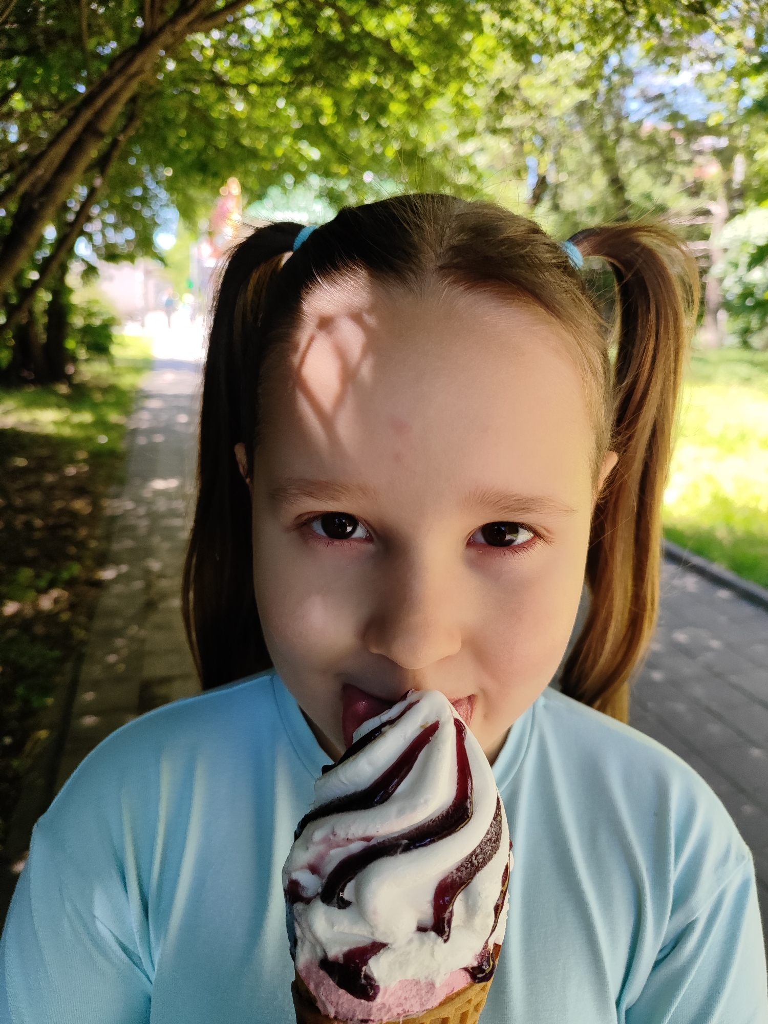 A close-up portrait features a young girl, approximately school-aged, looking directly at the viewer while licking an ice cream cone. She has light brown hair tied in pigtails with blue hair ties and is wearing a light blue t-shirt. Her dark brown eyes are wide and engaged.
The girl is holding a soft-serve ice cream cone, which appears to be a swirl of white (likely vanilla) and pink (possibly berry or strawberry) flavors, generously drizzled with a dark berry or chocolate syrup, all nestled in a waffle cone. Her tongue is visible as she licks the top of the ice cream.
The setting is outdoors in what appears to be a park or a tree-lined street in Krasnodar, Russia. The background is softly blurred with a bokeh effect, showing lush green foliage and tree branches overhead, creating a canopy. Dappled sunlight filters through the leaves, casting distinct shadows on the girl's forehead and the paved path visible behind her. Bright sunshine indicates a warm, clear day, further suggested by the activity of eating ice cream. The overall scene conveys a pleasant, summery atmosphere. No visible text is present in the image.