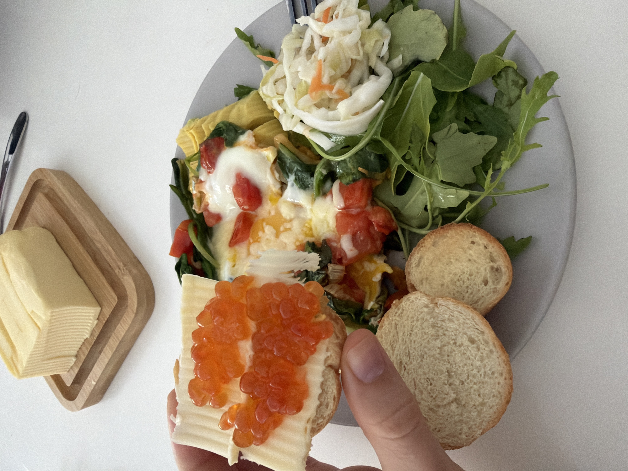 The image shows a meal on a white table. A light grey plate holds a mix of food, including a mound of shredded cabbage and carrots, leafy greens, a yellow omelet-like food item, and a mixture of spinach and chopped tomatoes topped with melted cheese or a white sauce. Two halves of a small loaf of bread rest next to the salad components. A hand holds a slice of bread covered with a thick layer of butter and orange caviar. A butter dish holding a block of butter sits to the left of the plate, accompanied by a silver butter knife. The location is unspecified, and there is no way to determine if the image was taken in Bat Yam, Israel.