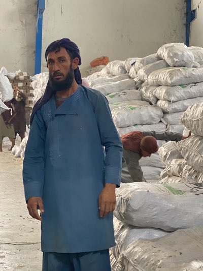 In a warehouse in Shurandam, Afghanistan, a man stands in the foreground, wearing a blue tunic and dark head covering. He has a beard and appears to be Middle Eastern. Behind him, a large stack of white sacks rises, reaching almost to the ceiling. A second man is partially visible, bent over and seemingly working with the sacks. In the background, other figures are visible amongst stacked boxes and sacks. The environment suggests a storage or distribution facility.
