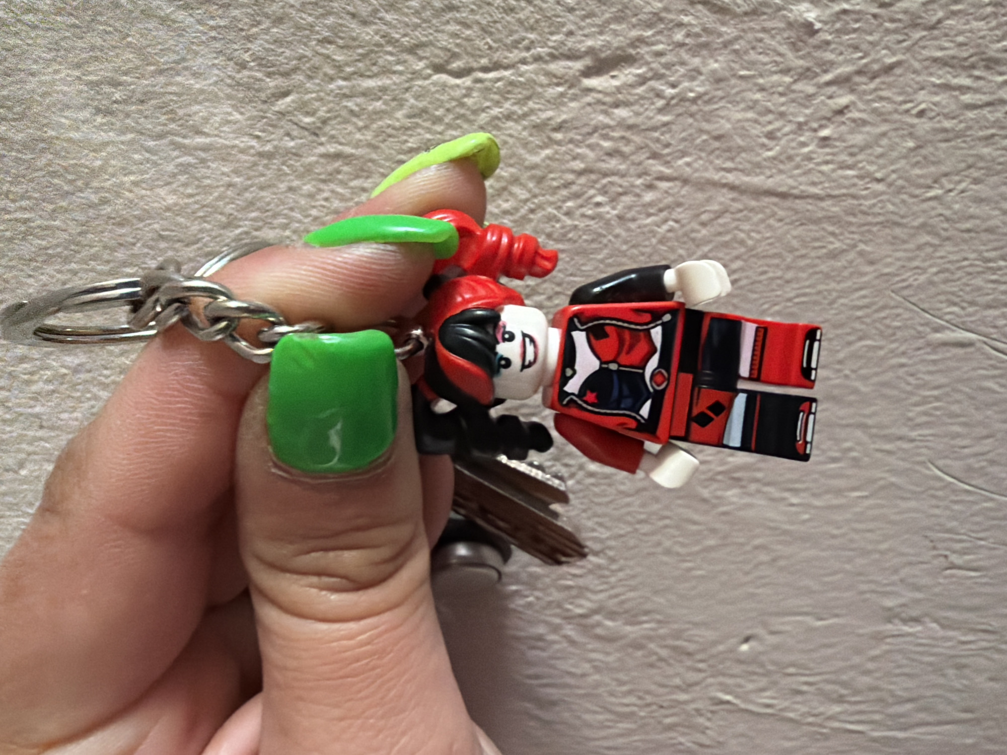 A close-up shot shows a hand holding a keyring with keys and a Lego Harley Quinn figure. The hand has three nails painted in different shades of green. The background is a textured wall with a neutral color.