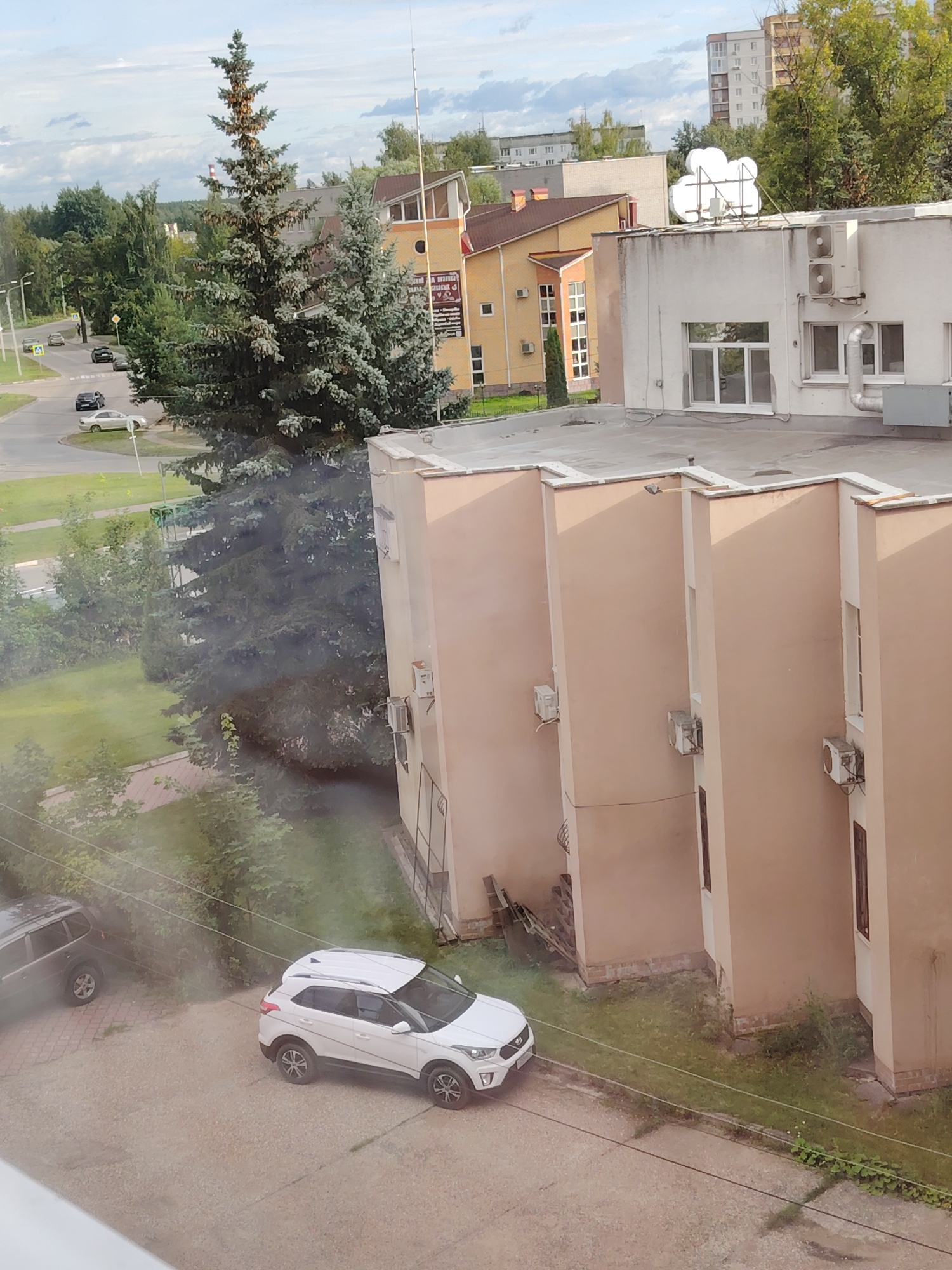 The image shows an aerial view of a street and buildings in Konakovo, Russia. In the foreground, a light-colored SUV is parked on a gravel surface. To the left, a dark-colored SUV is also parked. Behind these vehicles, a large evergreen tree dominates the mid-ground, with its branches extending towards the left side of the frame. A street with several cars driving on it is visible beyond the tree. In the background, a row of tan-colored buildings with multiple stories and windows is prominent. One of these buildings has air conditioning units mounted on its facade. Further in the distance, taller apartment buildings and more greenery can be seen under a cloudy sky. The scene appears to be a residential or commercial area.