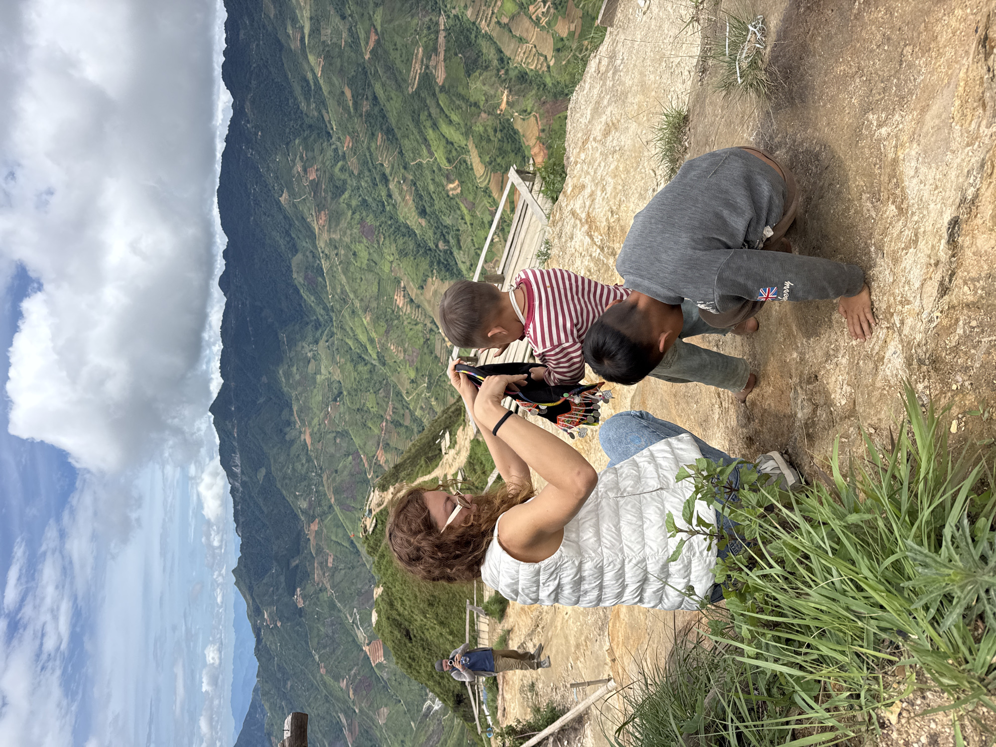 In Bắc Yên, Vietnam, two young boys and a woman are seen on a rocky ledge overlooking a vast valley with terraced hillsides. The woman, wearing a white puffer vest and jeans, is holding a colorful garment. One boy, in a red and white striped shirt, stands next to her. The other boy, in a gray sweatshirt, is crouched down, examining something on the ground. In the background, a man with a camera is visible. The sky is partly cloudy, and the overall scene suggests a scenic overlook with a natural landscape.