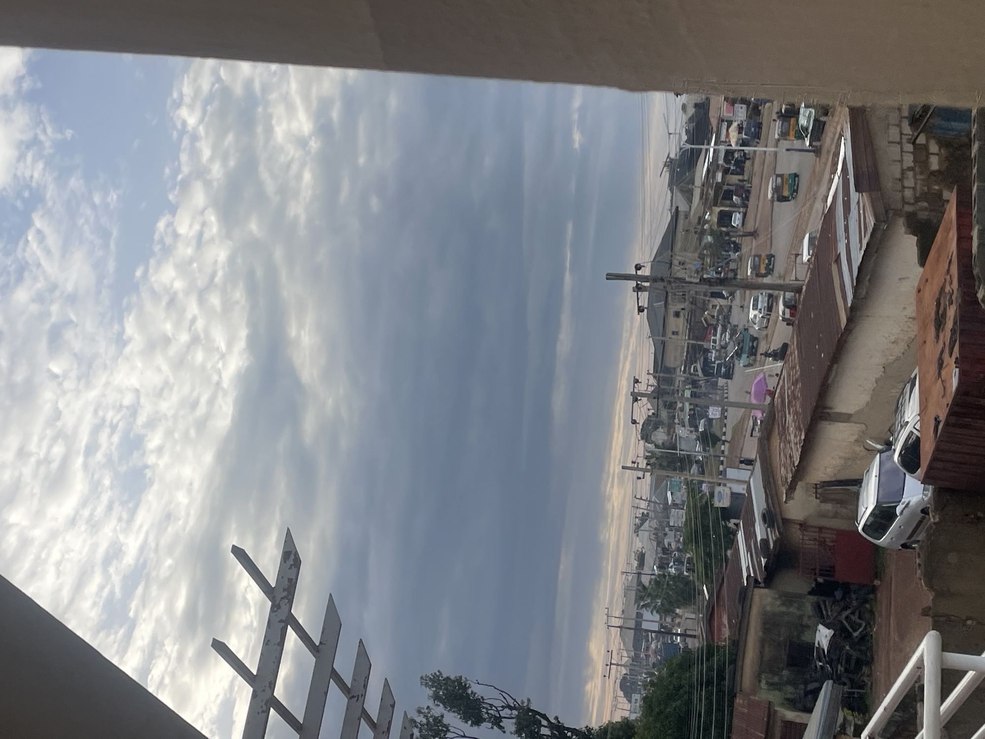 This is a photo taken from an elevated perspective, looking out over a street scene in Rafin Par, Nigeria. The image is oriented vertically, with the sky filling the left portion of the frame and the urban environment occupying the right. The sky is partly cloudy, with bright white clouds contrasting against a soft blue and grey backdrop, suggesting it's either early morning or late afternoon.
The street below is bustling with activity. Numerous vehicles, including cars, trucks, and what appear to be auto-rickshaws or tuk-tuks, are visible. The buildings are a mix of styles, with some having corrugated metal roofs and others appearing to be more permanent structures. There are utility poles and wires crisscrossing the scene, indicative of an active urban area. In the foreground, a white metal fence with pointed spikes is partially visible, framing the view. Some trees are also present, adding a touch of greenery to the otherwise built-up environment.
There is no discernible text visible in the image. The overall impression is of a typical day in a Nigerian town, with the weather appearing fair and the community engaged in its daily routines. The lighting suggests natural daylight, with no artificial illumination apparent.