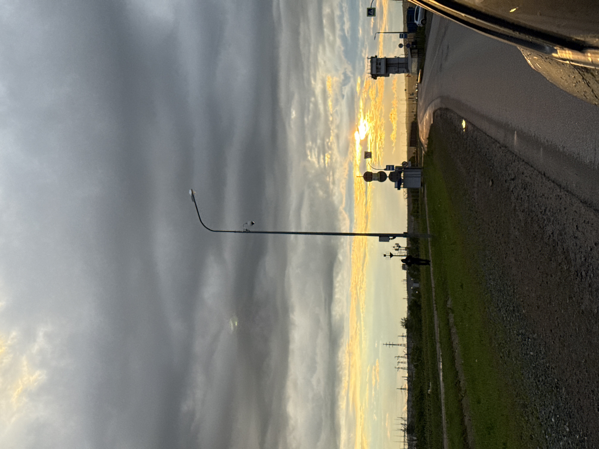 This dramatic image captures a roadside scene at either sunrise or sunset, viewed from inside a vehicle. The sky occupies a significant portion of the frame, dominated by heavy, dark grey clouds on the left that gradient into a breathtaking display of golden, orange, and bright white hues along the horizon to the right, indicating the position of the setting or rising sun.
In the foreground, a wide, wet asphalt road curves gently to the right, reflecting glints of light. To the right of the road, there's a gravel shoulder bordering a strip of green grass. A tall, slender streetlight or utility pole, equipped with what appears to be surveillance cameras, stands prominently on the grassy verge in the mid-ground. Further along the road and grass, the dark silhouette of a person is visible, walking.
The distant horizon is marked by the silhouettes of numerous utility poles or power line towers. A few indistinct roadside structures, possibly traffic signals or a small building, are also visible. The wet appearance of the road suggests recent rain. The overall mood is vast and somewhat serene, with a striking contrast between the dark, heavy clouds and the vibrant, warm glow of the horizon. No discernible text is visible in the image.
