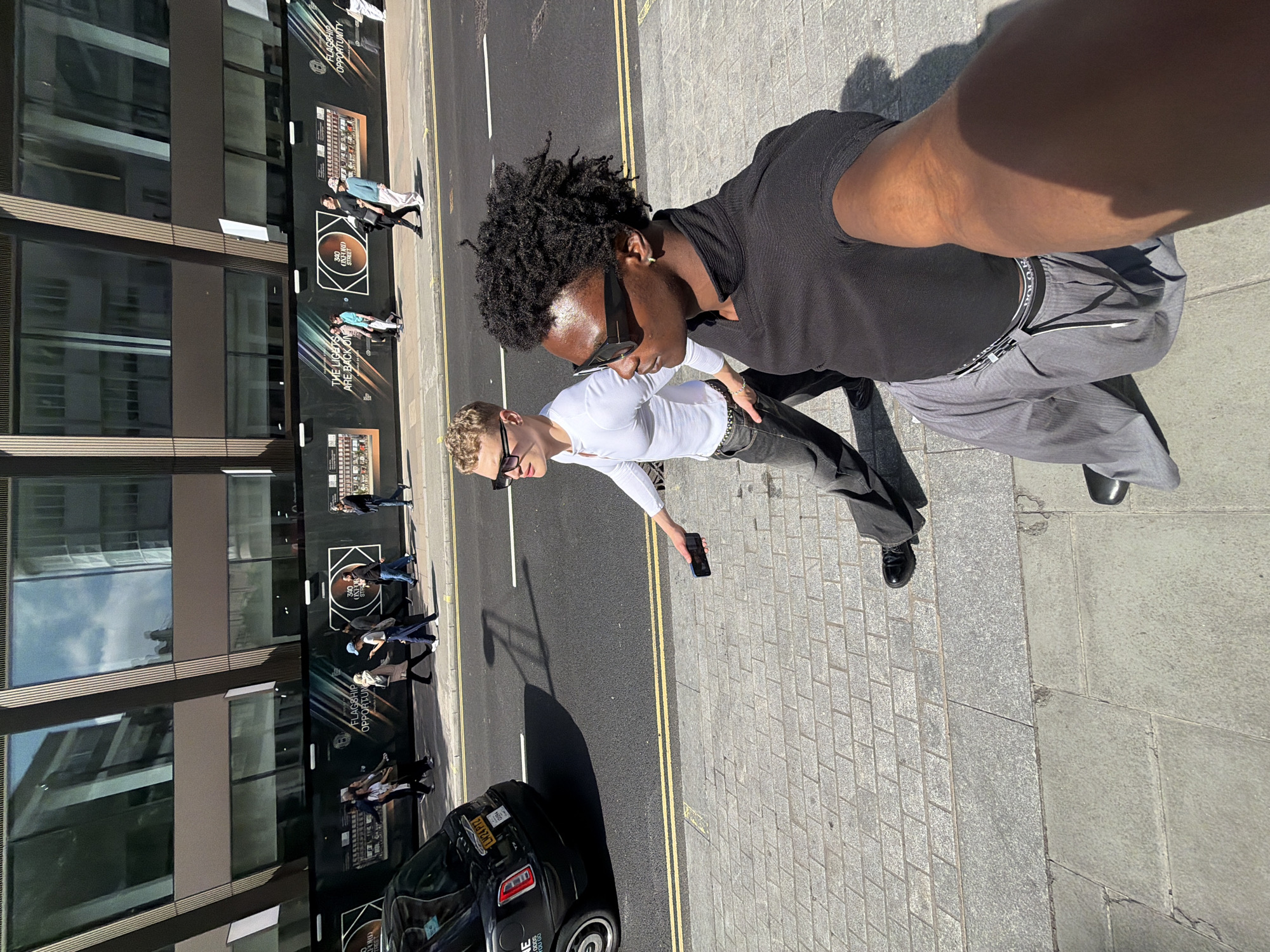 Here's a detailed description of the image:
**Overall Scene:** The image appears to be a selfie taken on a sunny day on a street in London. The photographer is positioned on a sidewalk next to a road with double yellow lines. The background features a modern building with large windows, partially covered by construction hoarding. People are walking on the opposite sidewalk.
**People:**
* **Photographer:** A person with dark skin, wearing a black t-shirt and grey pants with visible underwear band.
* **Second Person:** A lighter-skinned individual in a long-sleeved white shirt and dark pants is walking beside the photographer. They have sunglasses and are holding a smartphone.
**Objects:**
* **Buildings:** A modern building with large windows and construction signage is visible in the background.
* **Car:** A black vehicle, possibly a Range Rover, is parked on the street.
* **Sidewalk:** Grey paved sidewalk.
* **Road:** An asphalt road with double yellow lines.
**Actions:**
* The photographer is taking a selfie.
* The second person is walking and holding a smartphone.
* Other people are walking on the opposite sidewalk.
**Environment:**
* The lighting suggests a bright, sunny day.
* The setting is urban, likely a commercial area with a mix of buildings and traffic.
**Location in the city of London, United Kingdom:**
The construction hoarding features the text "340 Oxford Street". Therefore, the image location is Oxford Street.