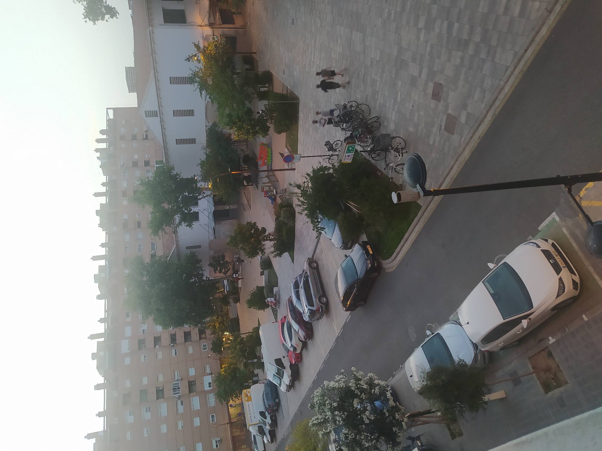 The image shows a street view in a city. The scene is an outdoor environment with buildings, cars, people, and bicycles. In the background, there are multi-story buildings with many windows. In the foreground, a street or parking lot is visible with several cars parked. Some of these cars are white, while others are dark-colored. There is also a white van. A few people are walking on the sidewalk or in a plaza area. Several bicycles are parked together. Trees and bushes are scattered throughout the scene, and there is a lamppost. The sky is overcast, suggesting it might be early morning, late afternoon, or a cloudy day.