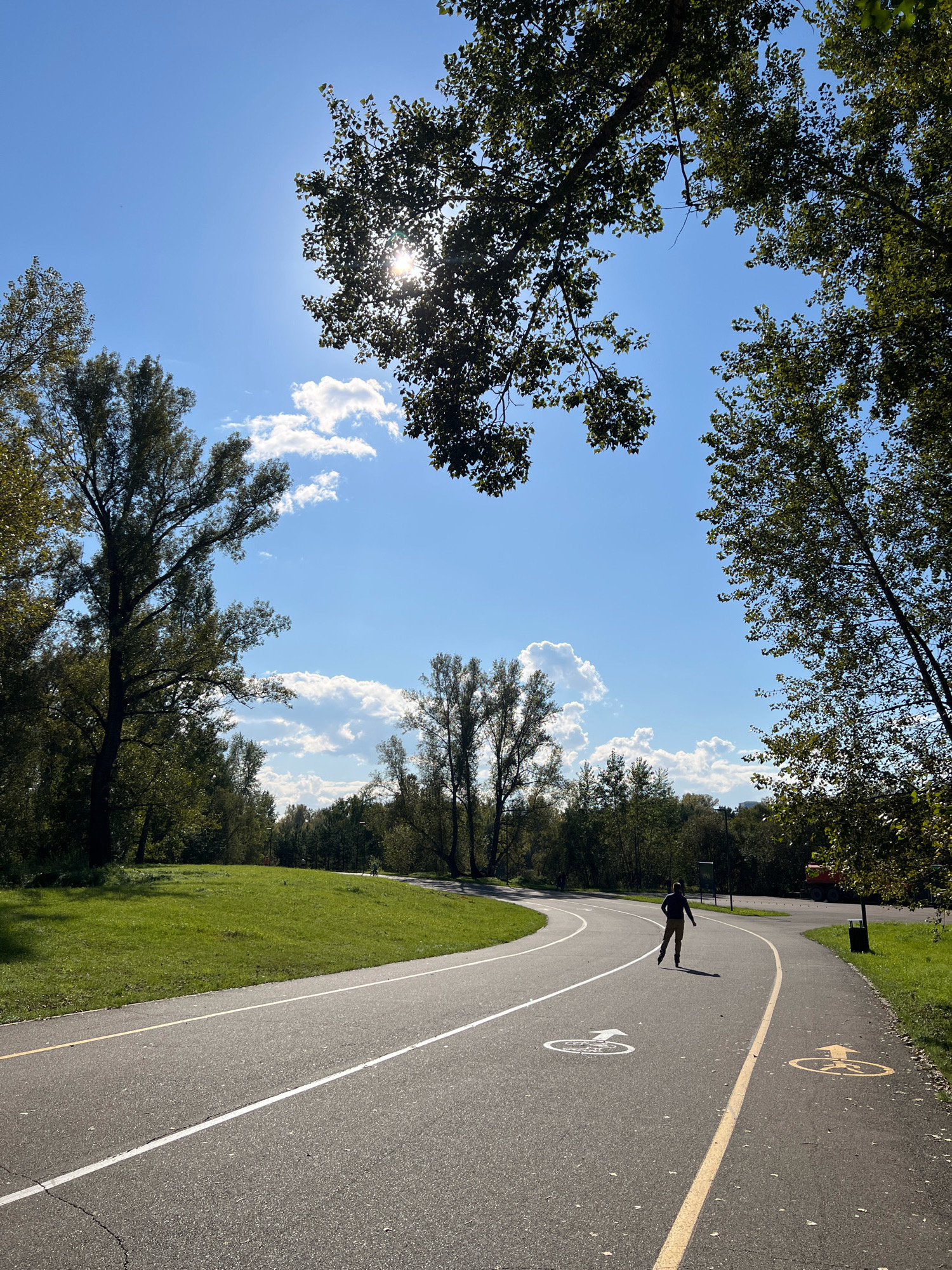 The image depicts a park scene with a paved pathway winding through lush green grass and trees. The sky is a bright blue with scattered white clouds, and the sun is visible through the leaves of trees, creating a dappled light effect. A person is rollerblading on the pathway, casting a shadow. The pathway has painted markings, including arrows and symbols indicating cycling and walking lanes. In the background, more trees and a distant orange vehicle are visible. The scene conveys a sense of outdoor recreation and a pleasant day in a park. The location is identified as a park in the city of Krasnoyarsk, Russia.