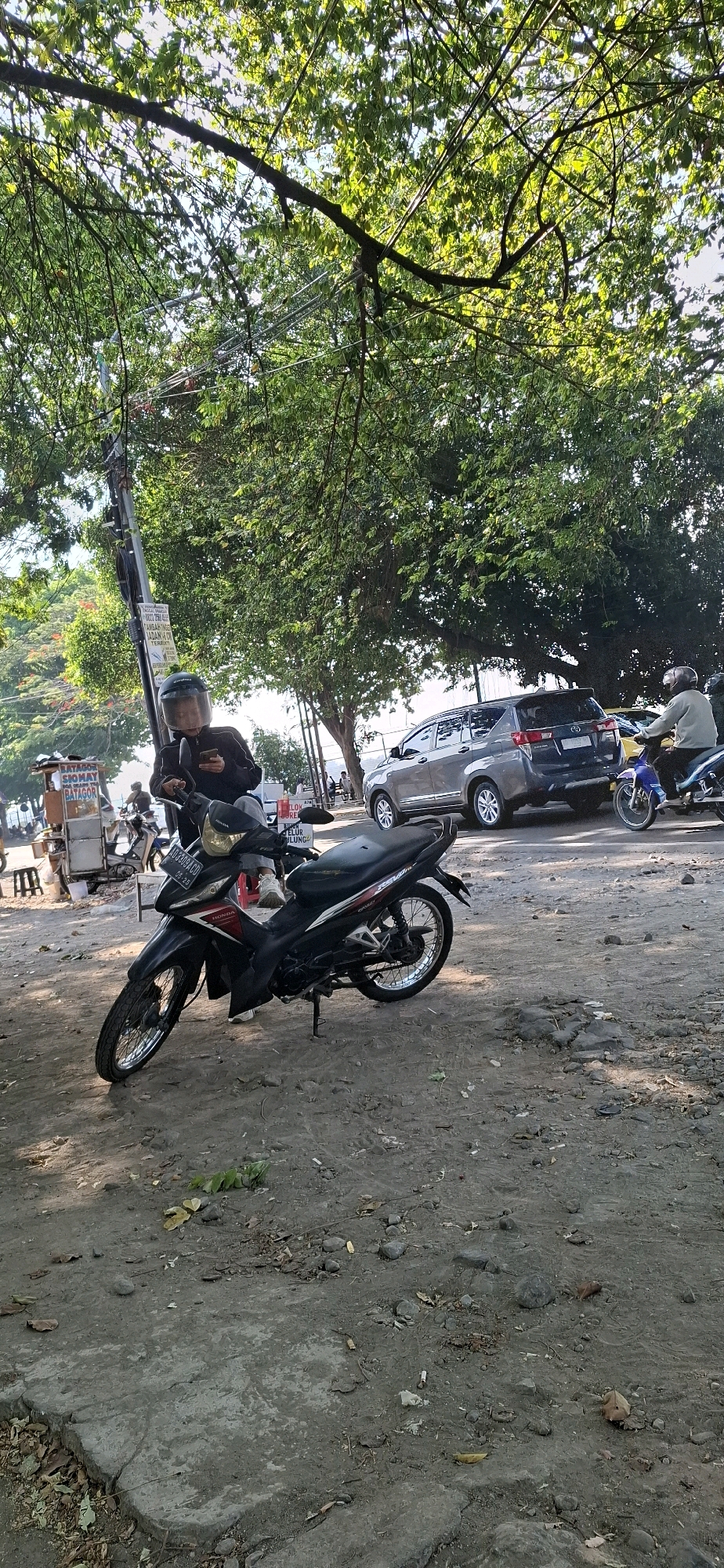 This image captures a street scene in Desa Ngesrep, Indonesia, under the dappled shade of lush green trees. In the foreground, a black and red Honda motorcycle is parked on a dirt and concrete ground scattered with leaves and small rocks. A person wearing a black jacket and a helmet with a clear visor is seated on the motorcycle, looking at their phone. Behind the motorcycle, a dark grey SUV is parked, and a person on a blue motorcycle is visible to the right. In the background, a food cart with signage in Indonesian, including "SIOMAY" and "BATAGOR", is set up on the left. The environment suggests a warm, possibly humid day with sunlight filtering through the canopy. The overall impression is a snapshot of daily life in a tropical urban or semi-urban setting.