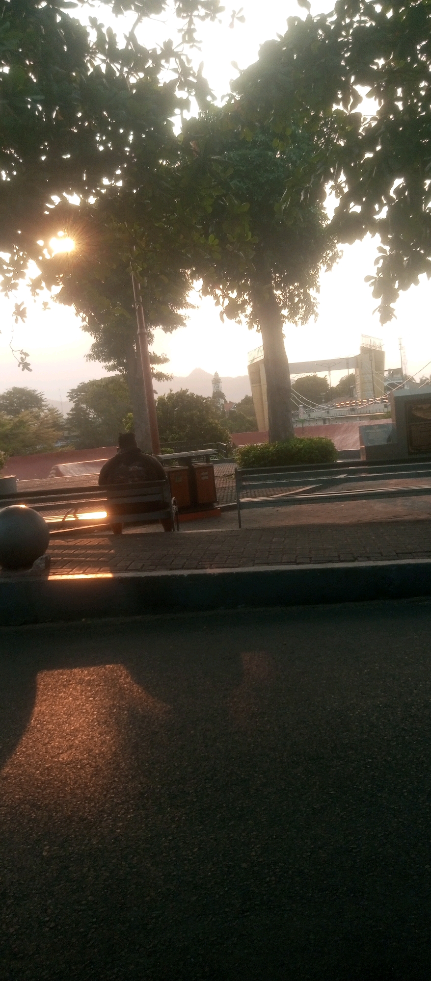 The image depicts a street scene in Kediri City, Indonesia, during a sunrise. The sun, with visible rays, is positioned behind the trees in the upper left quadrant. In the foreground, a dark asphalt road is visible, with a patch of sunlight reflecting on it. To the left, a large grey spherical object is partially visible. Behind this, a park bench with a dark figure sitting on it is present, illuminated by the setting sun's rays. Orange waste bins are situated behind the bench. The middle ground features a paved walkway with a low wall. In the background, a bridge structure with yellow pillars and suspension cables is visible. Further in the distance, trees and mountains are present, with a white tower and spire visible on a distant hill. The overall environment suggests an outdoor public space, possibly a park or promenade, in Kediri City.