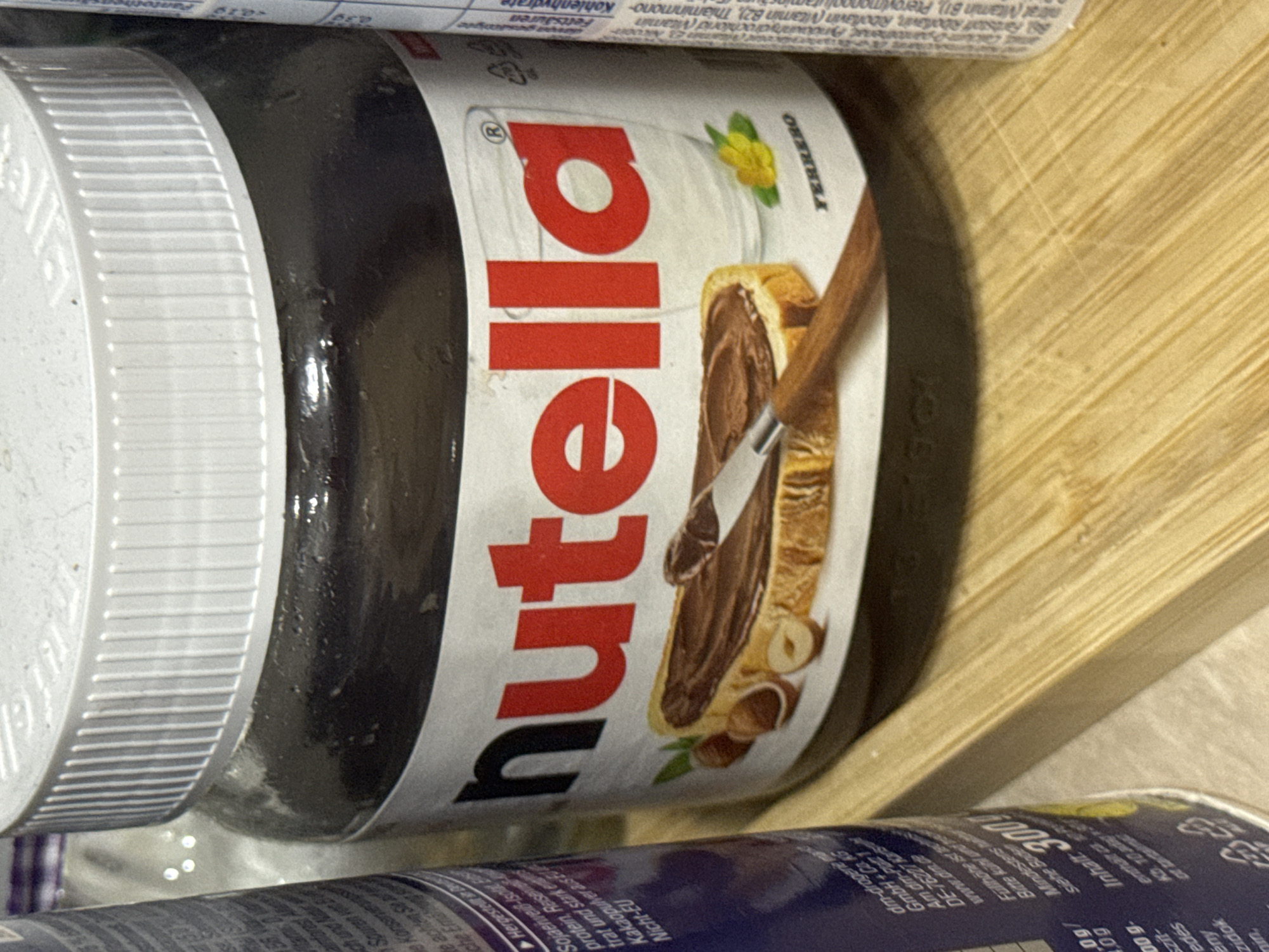 The image shows a close-up of a jar of Nutella, a popular hazelnut spread. The jar is mostly filled with the dark brown spread and has the iconic Nutella logo prominently displayed on its label. The label also features an illustration of toast with Nutella spread and hazelnuts. Next to the Nutella jar, parts of other products or packaging are visible, including what appears to be a blue bottle on the left and a white cylindrical object with text on the right, possibly another food product or supplement. The items are placed on a light-colored, textured surface that resembles wood. The lighting suggests an indoor environment. There is no indication of the city of Tbilisi, Georgia, or any specific location within it present in the image.