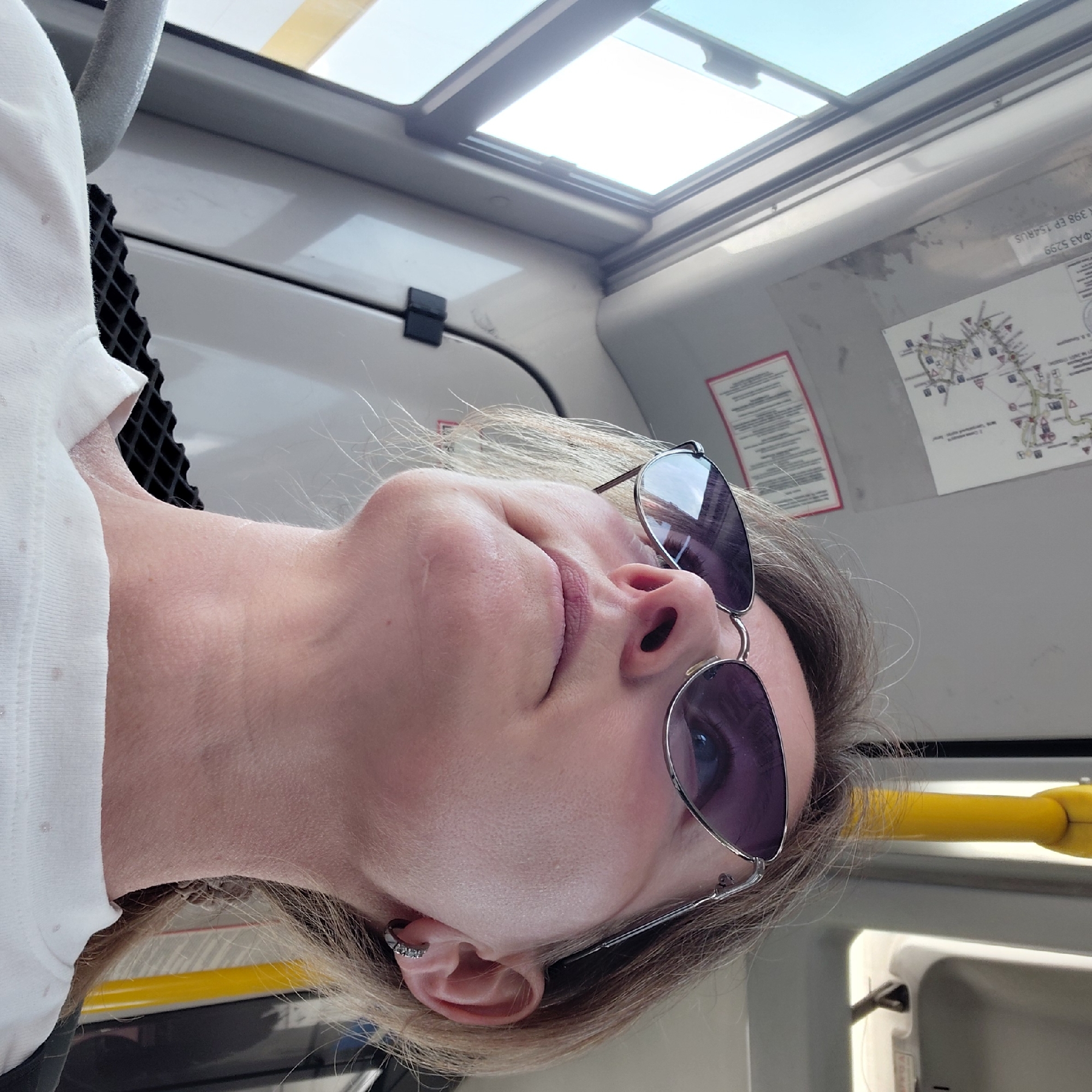 An adult Caucasian female with blonde hair pulled back is photographed from a low angle while looking upwards and to the right. She is wearing silver-rimmed sunglasses with purple lenses and a white t-shirt. A small, silver hoop earring with three clear stones is visible in her left ear. The background suggests she is inside a public transportation vehicle, likely a bus or train, with a yellow handrail, a map, and window frames visible. The scene is brightly lit by sunlight coming from the left. There is no indication of the specific location within Novosibirsk, Russia.