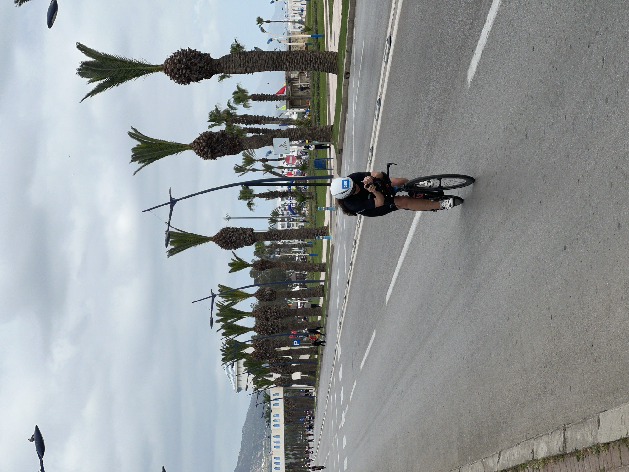 The image shows a cyclist on a road in M'Diq, Morocco. The cyclist, wearing a white helmet with the number 200 on it, is riding a black racing bike. The road is lined with palm trees and streetlights. Other cyclists can be seen further down the road. The sky is overcast. In the background, there are buildings and a mountain.