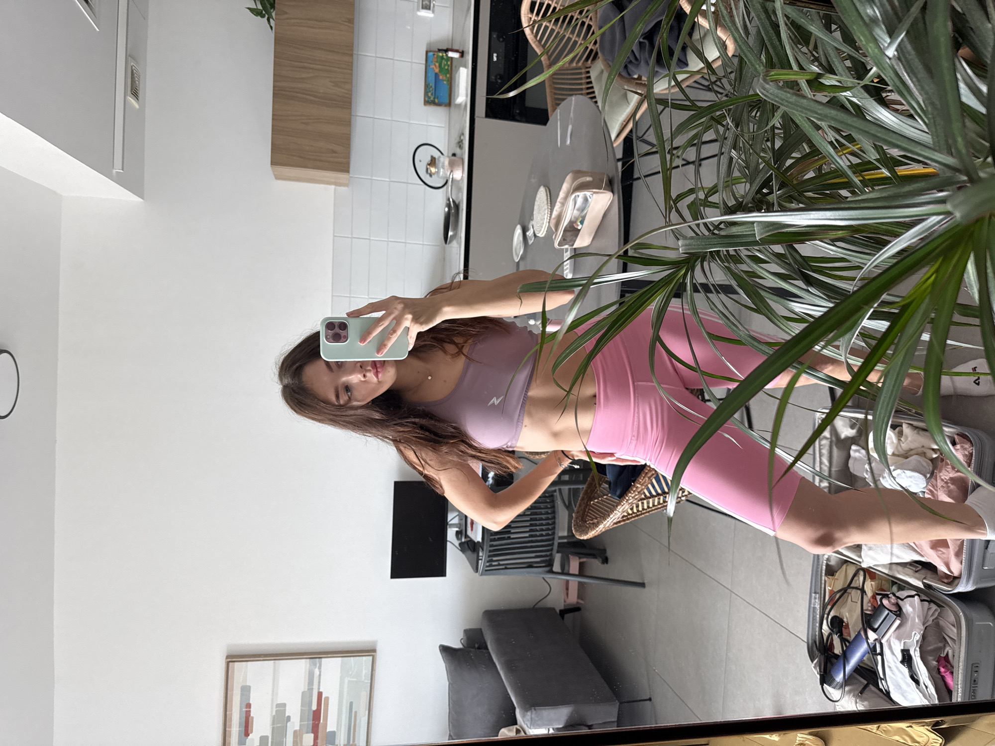 A young woman is captured taking a mirror selfie in what appears to be a modern apartment in Tel-Aviv, Israel. She is seen wearing a light purple athletic bra top with a white "N" (or similar) logo on the front, paired with pink high-waisted biker shorts and white socks. She holds a light green smartphone in her right hand to take the photo, while her left hand is placed on her hip. Her long brown hair flows down her shoulders.
The reflection in the mirror reveals parts of the apartment's interior. To the woman's left, a living area is visible with a white wall, a black flat-screen television on a stand, a gray sofa with dark pillows, and a framed abstract painting on the wall. To her right, a kitchen area features white tiled walls, light wooden upper cabinets, and a kitchen counter or island displaying various items such as a small pan, decorative objects, a light pink makeup bag, and brushes. A wicker chair is also present in this section of the room.
A large potted plant with long green leaves partially obscures the scene on the right, closer to the mirror. On the floor, an open light gray suitcase is filled with clothes and what appears to be a blue hair dryer or curling iron, hinting at travel or recent unpacking. The bright, natural lighting suggests it is daytime. The image conveys a casual, personal moment within a contemporary home setting.