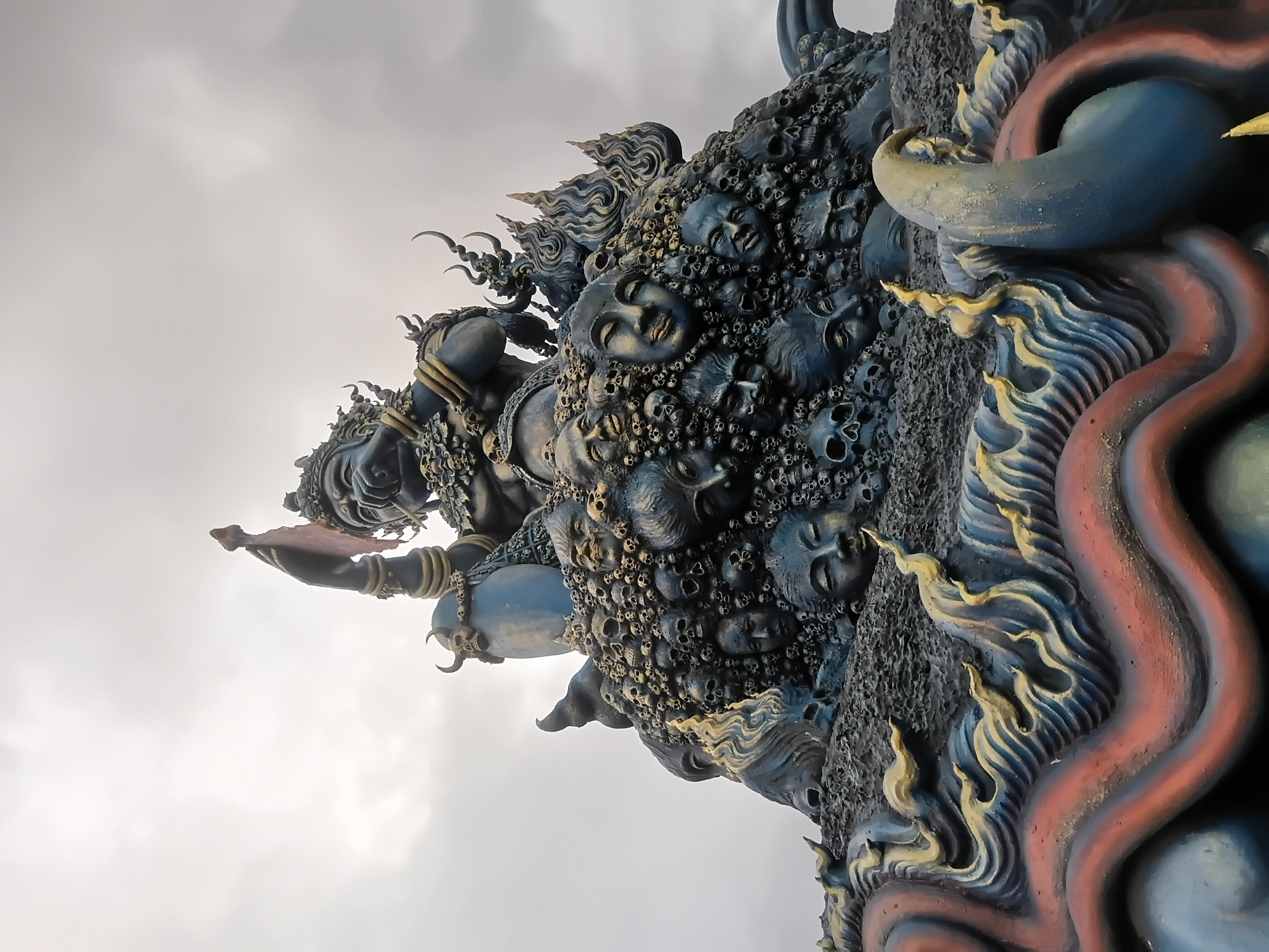 A low-angle, full shot shows the upper portion of a dark, imposing sculpture against a bright, overcast sky. The sculpture is composed of numerous dark, blue-toned human faces and skulls densely packed together, forming a mound. At the very top, a larger, dark figure with golden armbands and jewelry sits, holding a tattered red flag. The figure has a horned headdress and a stern expression. The sculpture's base features swirling, flame-like forms in red, blue, and gold, suggesting movement and intensity. This sculpture is located at Wat Rong Khun, also known as the White Temple, in Chiang Rai City Municipality, Thailand.
