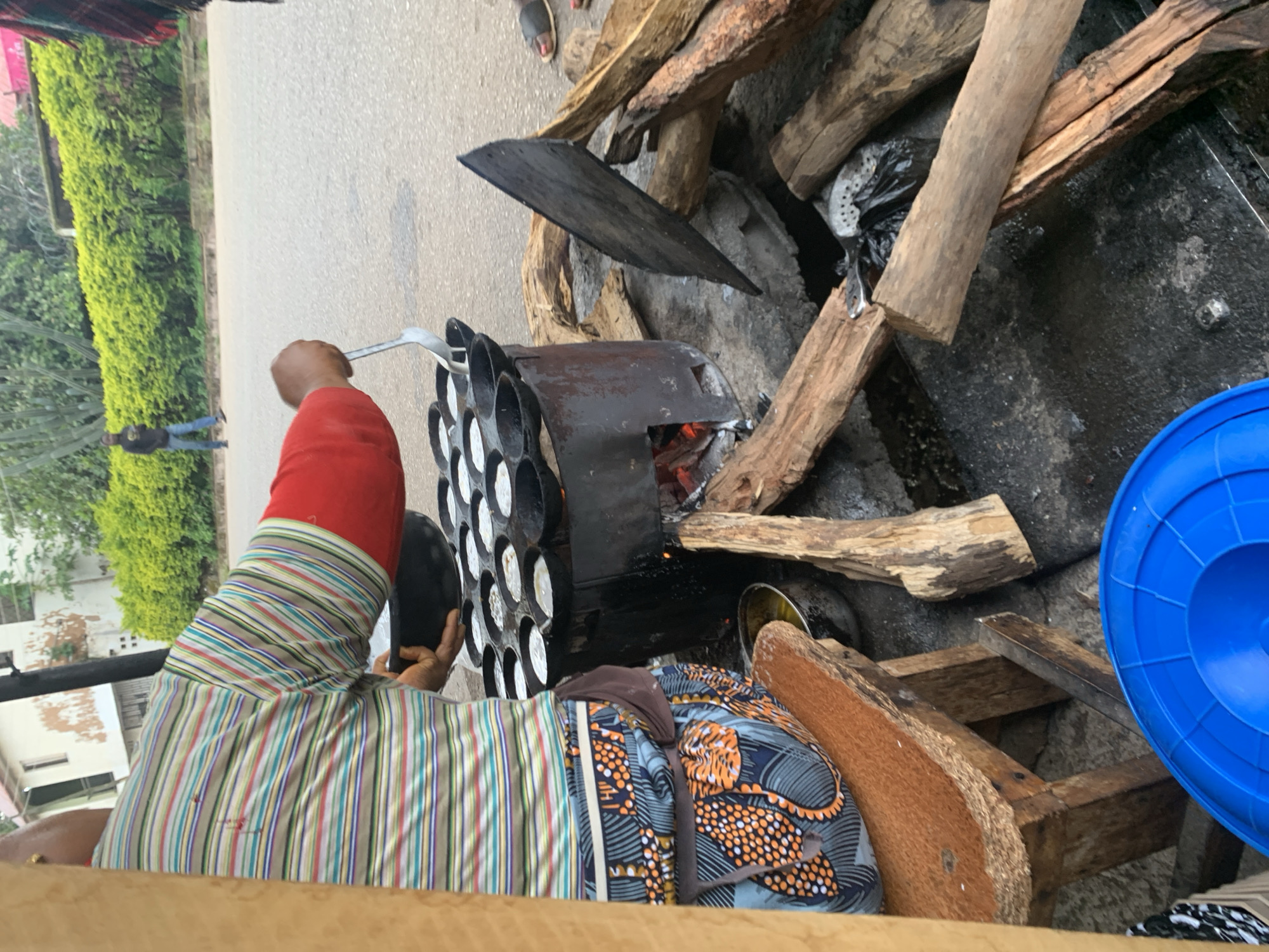 An outdoor scene in Jos, Nigeria, features a woman preparing food over a charcoal fire. She is wearing a horizontally striped long-sleeved shirt and a patterned skirt. The woman is using a ladle to pour batter into a multi-cavity cooking pan placed on a makeshift stove. Nearby, several logs of wood are stacked, some of which are burning in the stove. A large blue plastic container is visible in the foreground. In the background, a man walks on a paved road in front of a hedge and a building. The ground is wet and appears to be asphalt.