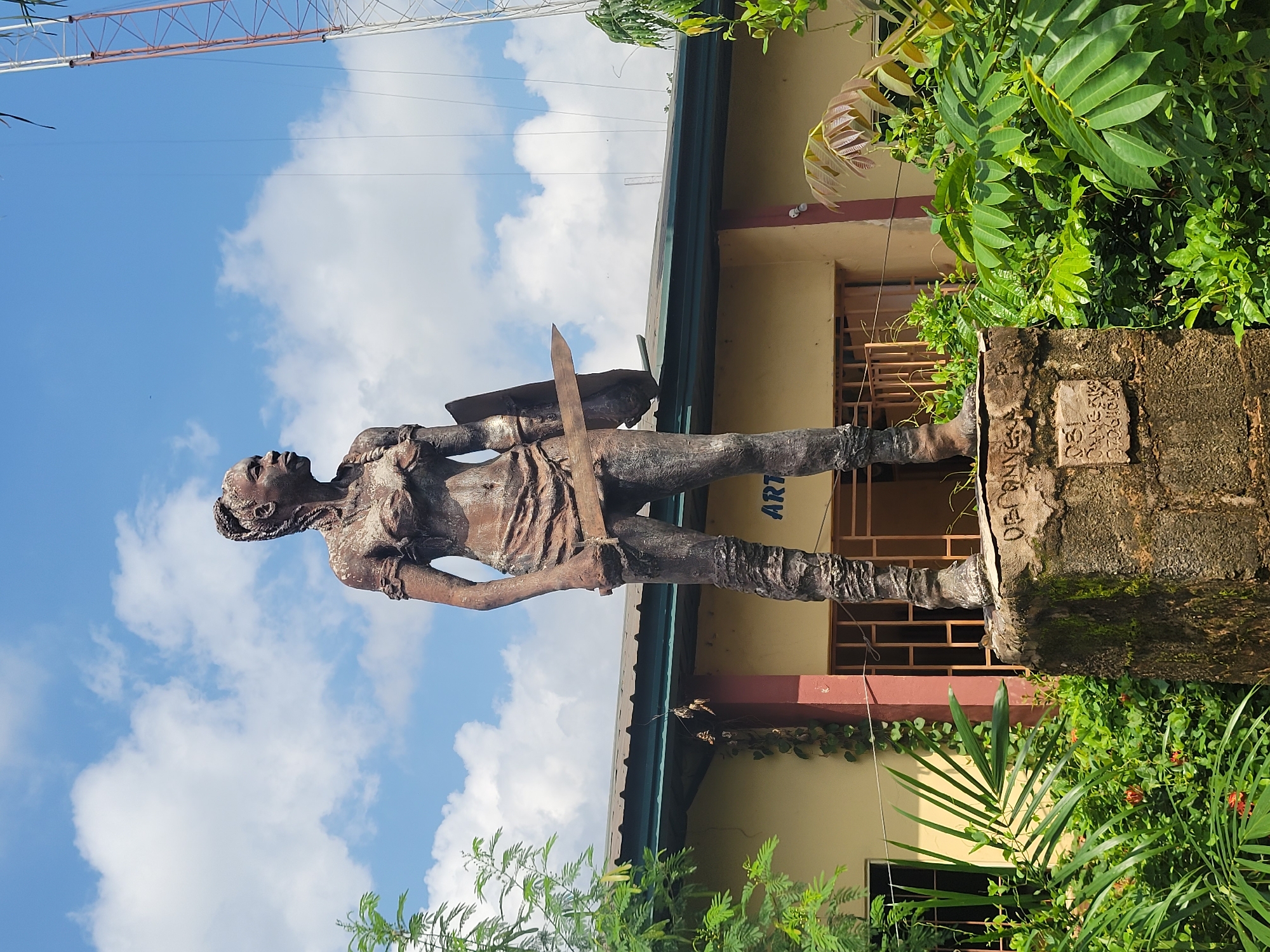 This media file depicts a statue of a person holding a sword and a shield, positioned in front of a yellow building with red trim. The statue appears to be made of bronze and is weathered, suggesting it is an outdoor monument. Green foliage surrounds the base of the statue and climbs the side of the building. The sky in the background is blue with scattered white clouds, indicating fair weather. A tall structure, possibly a radio tower or antenna, is visible in the upper left corner of the image. The building has windows with metal grates.
Visible text on the base of the statue or a plaque is partially obscured but appears to include what might be a name or inscription. The overall scene suggests a public space in a tropical or sub-tropical environment, likely outdoors during the daytime. The statue's pose and the items it holds imply a historical or cultural significance, possibly representing a warrior or a figure of importance. The rotation of the image suggests it was taken with a phone and is currently oriented sideways.